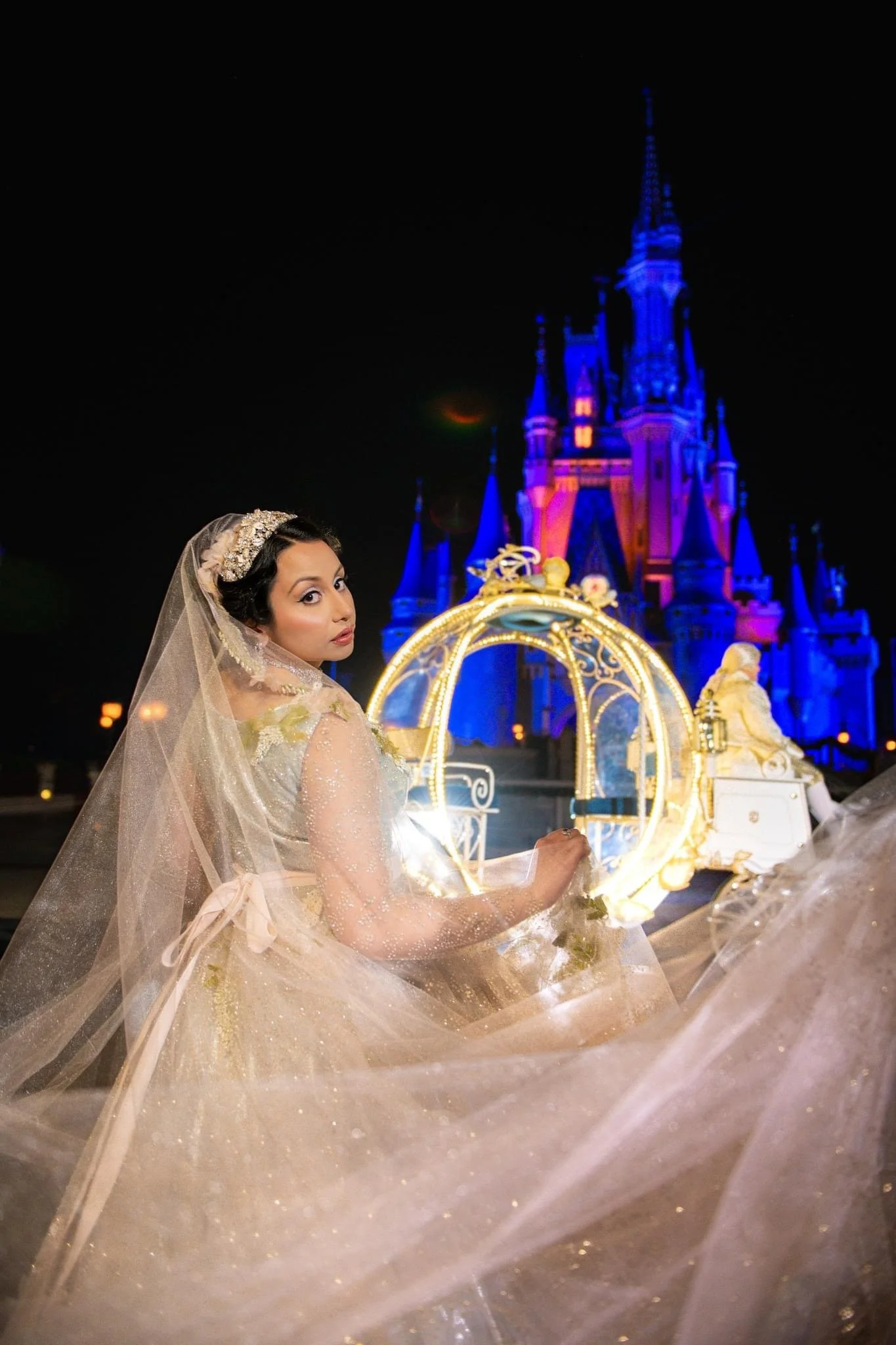 A woman in an Indian inspired wedding dress and veil outdoors at night, with a lit Cinderella carriage and a Disney castle illuminated in the background.