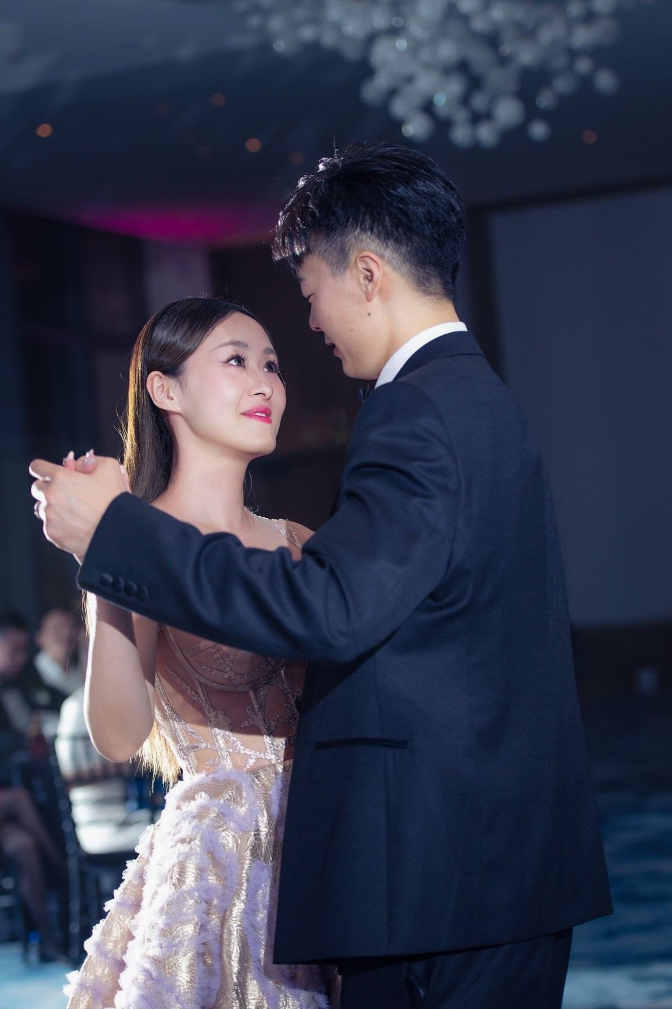 A young man and woman are dancing closely in formal attire, with the woman looking emotional and the man smiling, in a decorated indoor venue.