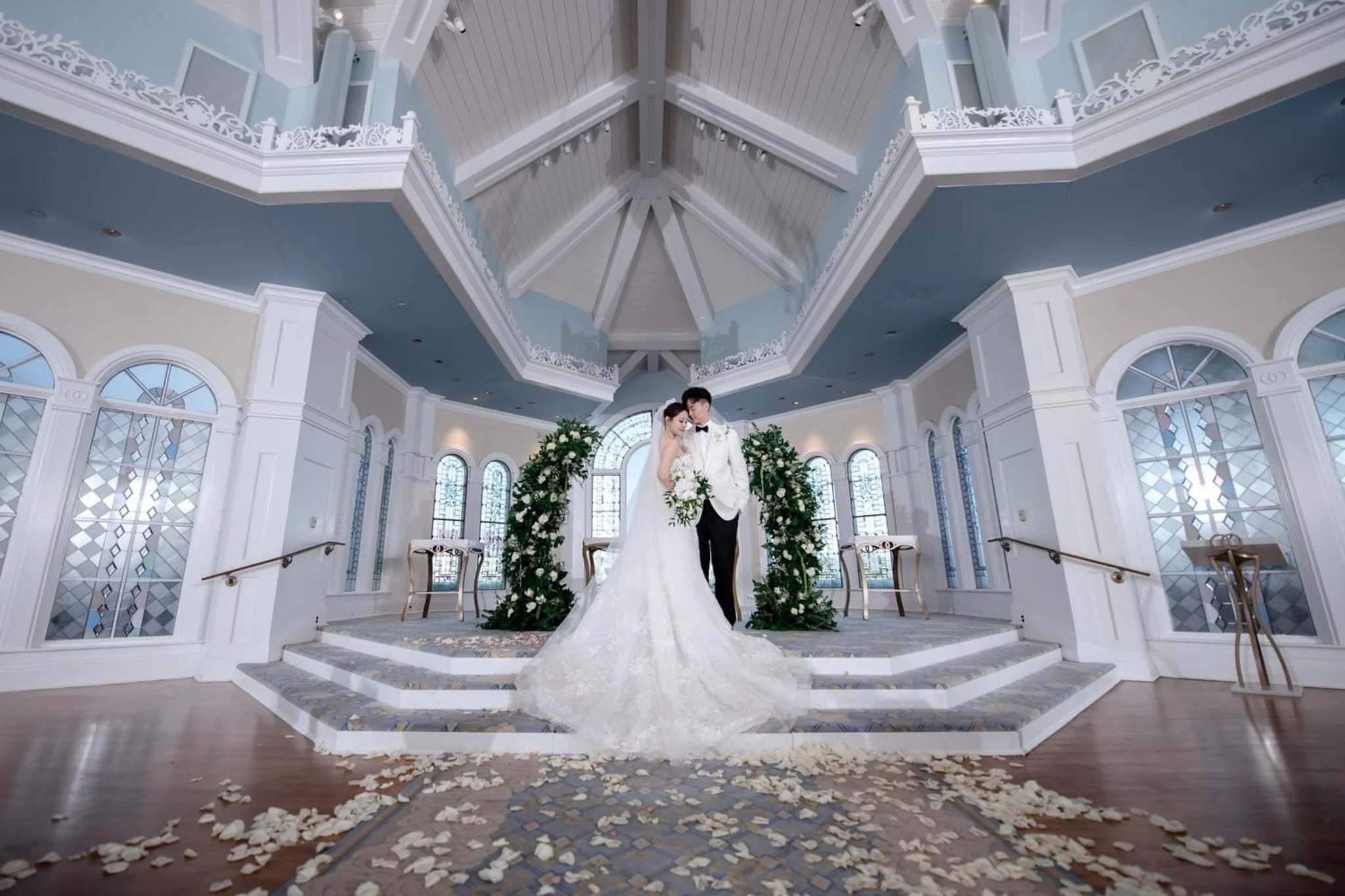 A bride and groom standing together on a staircase in at Disney's Wedding Pavilion with floral archways and large windows.