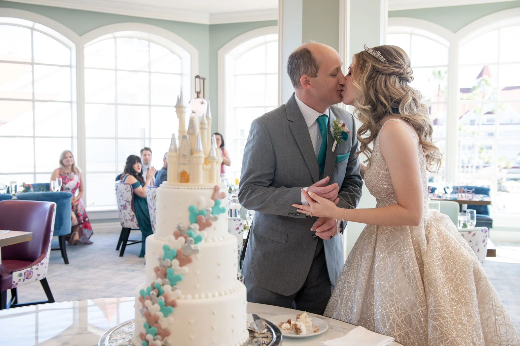 A bride and groom kiss at their wedding reception, with a wedding cake in the foreground and guests clapping in the background.