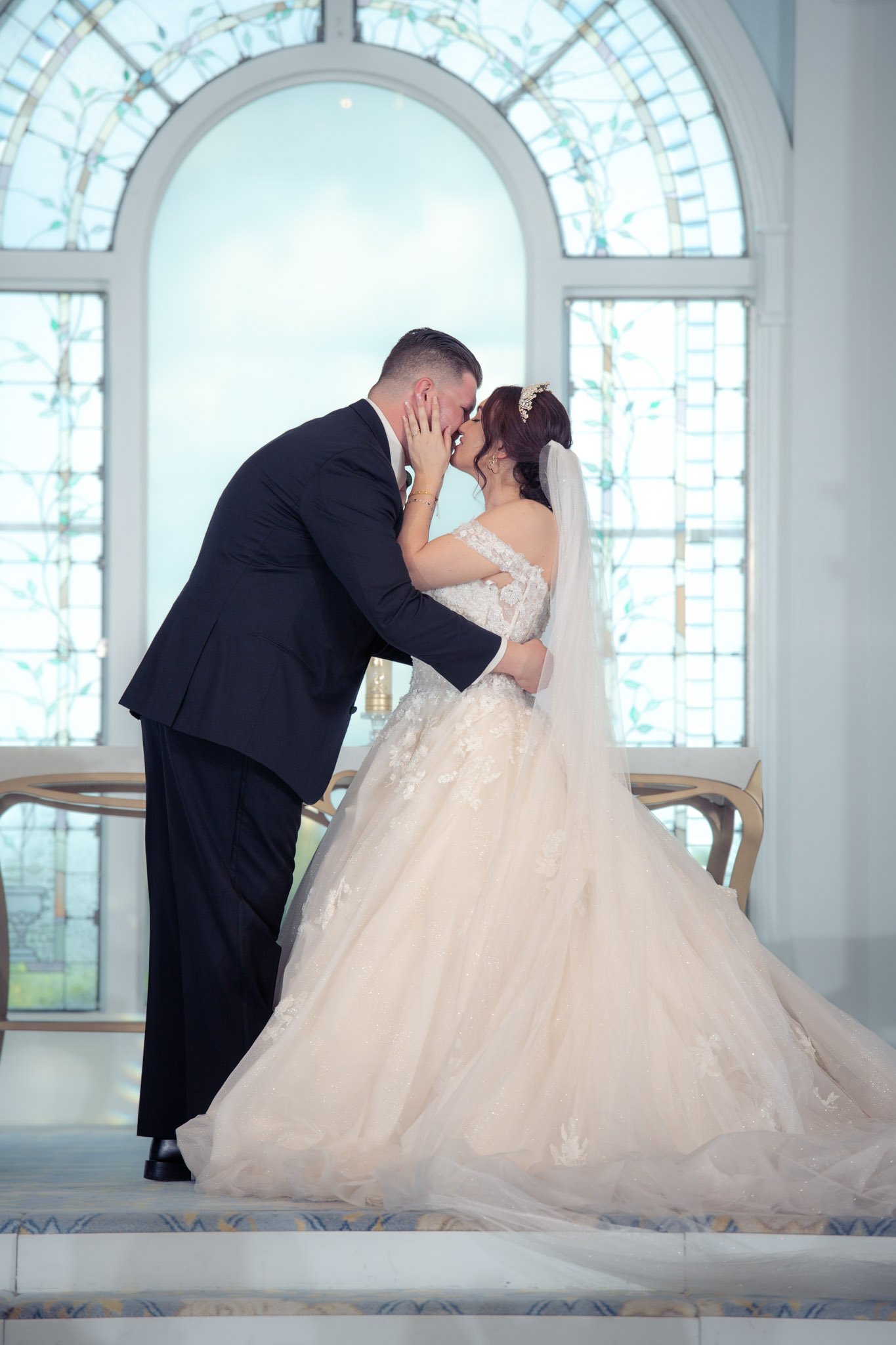 A bride and groom share a kiss during their wedding ceremony inside Disney's Wedding Pavilion with stained glass windows in the background.
