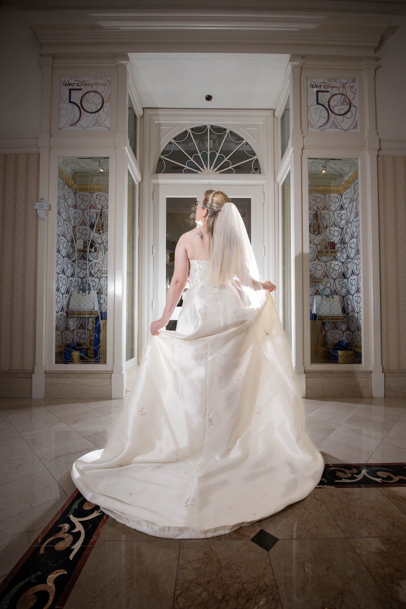 A bride in a white wedding dress and veil standing in an elegant hotel lobby, back facing the camera, holding the train of her dress.