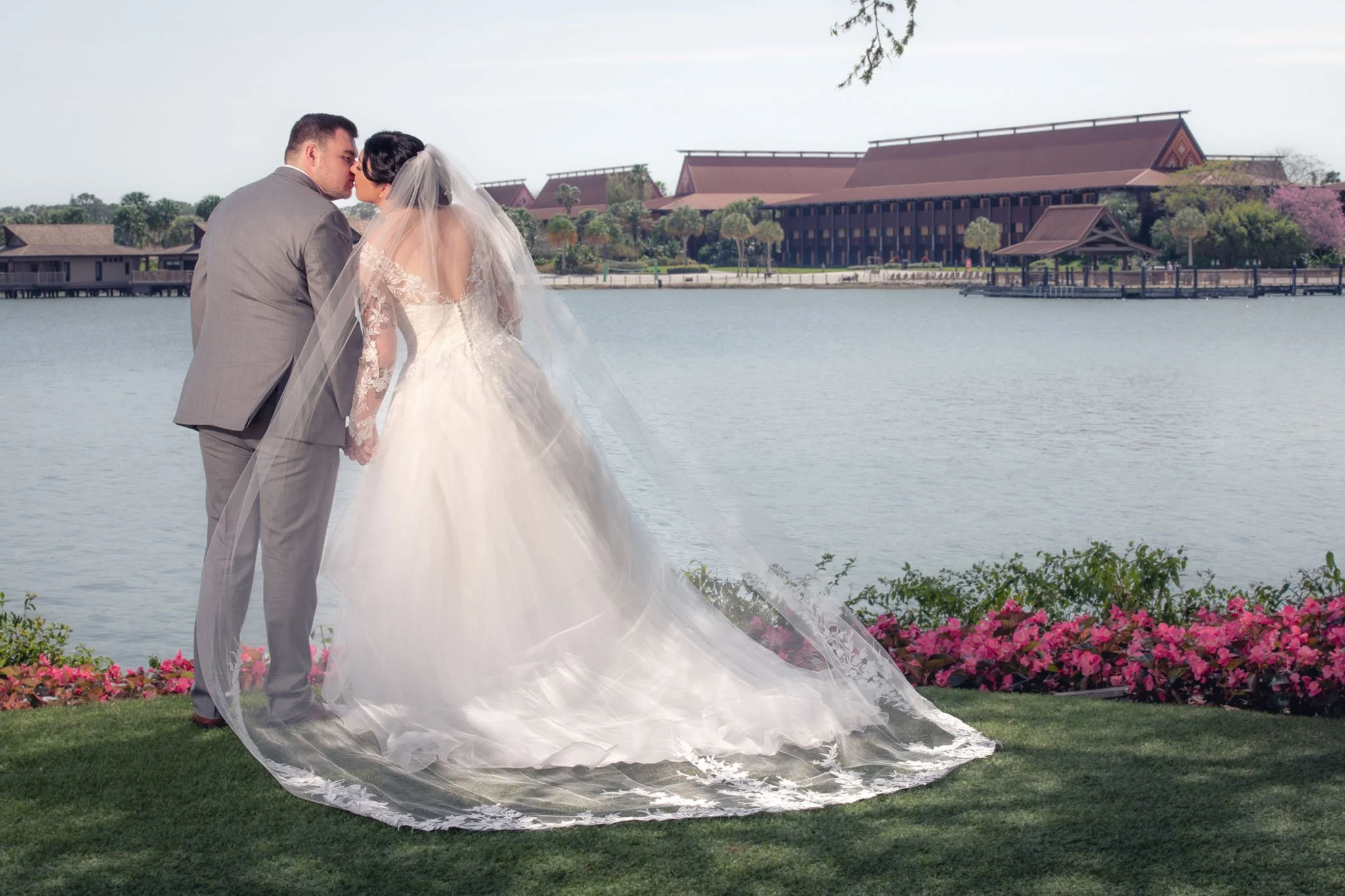 A bride and groom dressed in wedding attire, standing by a lake with a Disney's Polynesian Resort in the background, ponds, and pink flowers in the foreground.