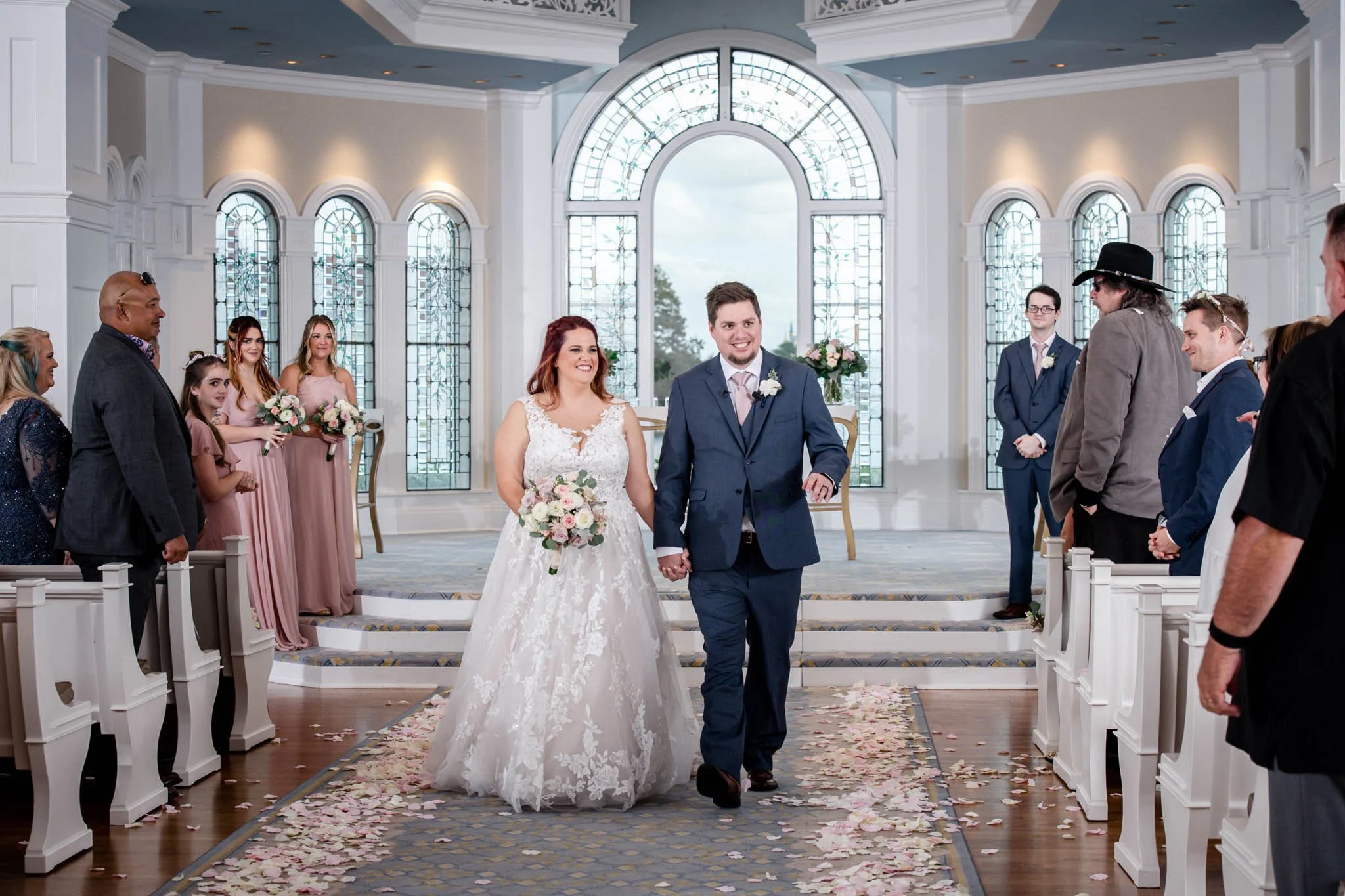 A bride and groom walking down the aisle hand in hand at Disney's Wedding Pavilion, surrounded by their wedding party and guests, with flower petals on the floor.
