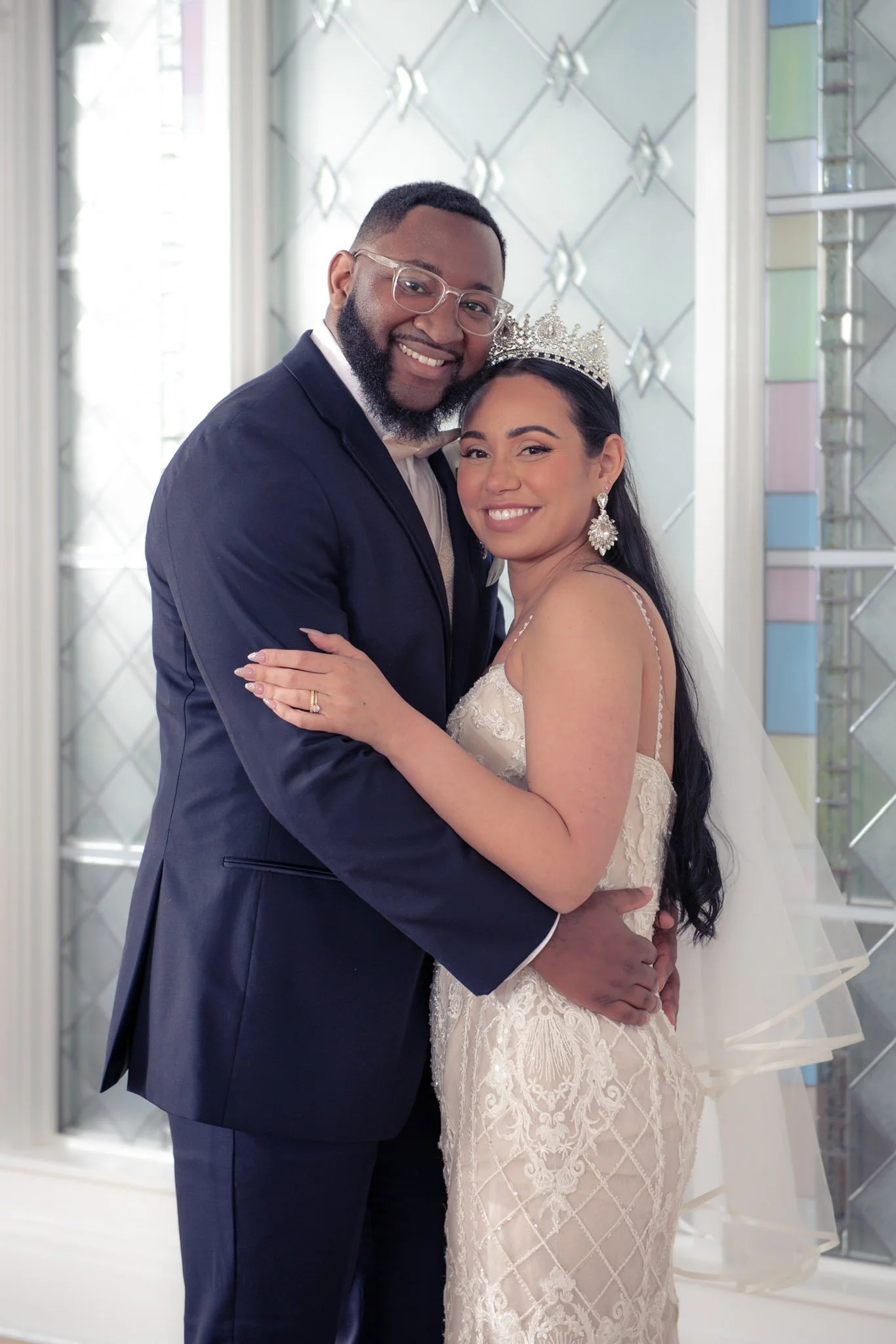 A happy couple, a man in a tuxedo and a woman in a wedding dress with a tiara and veil, embracing and smiling, in front of a decorative mirrored wall.