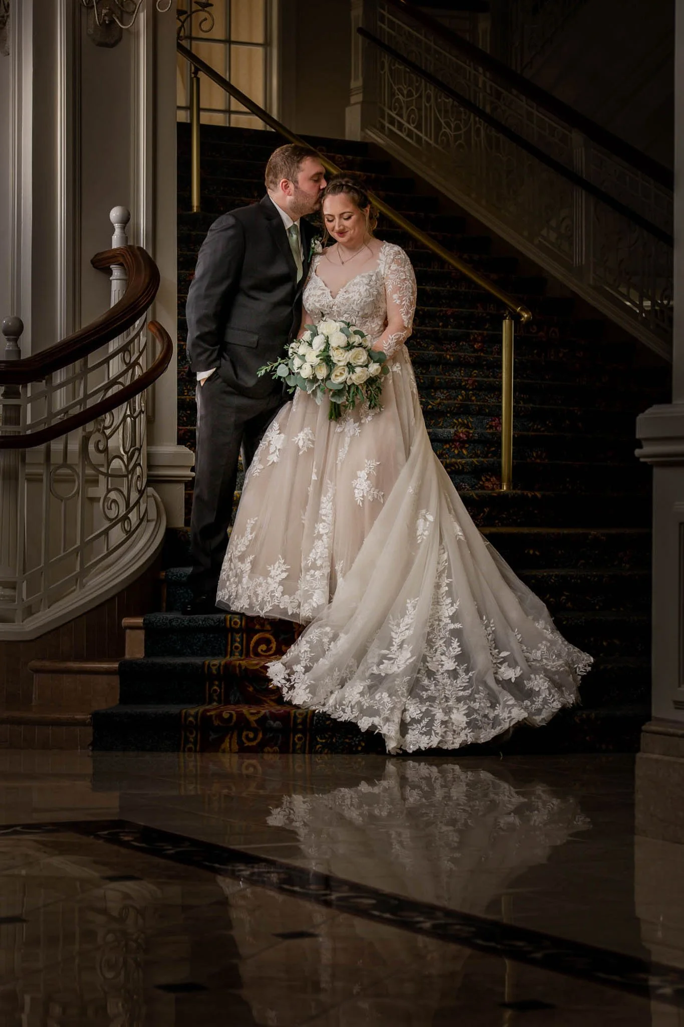 A bride and groom standing on a staircase, with the groom kissing the bride's forehead. The bride is wearing a lace wedding gown and holding a bouquet of white roses and greenery.