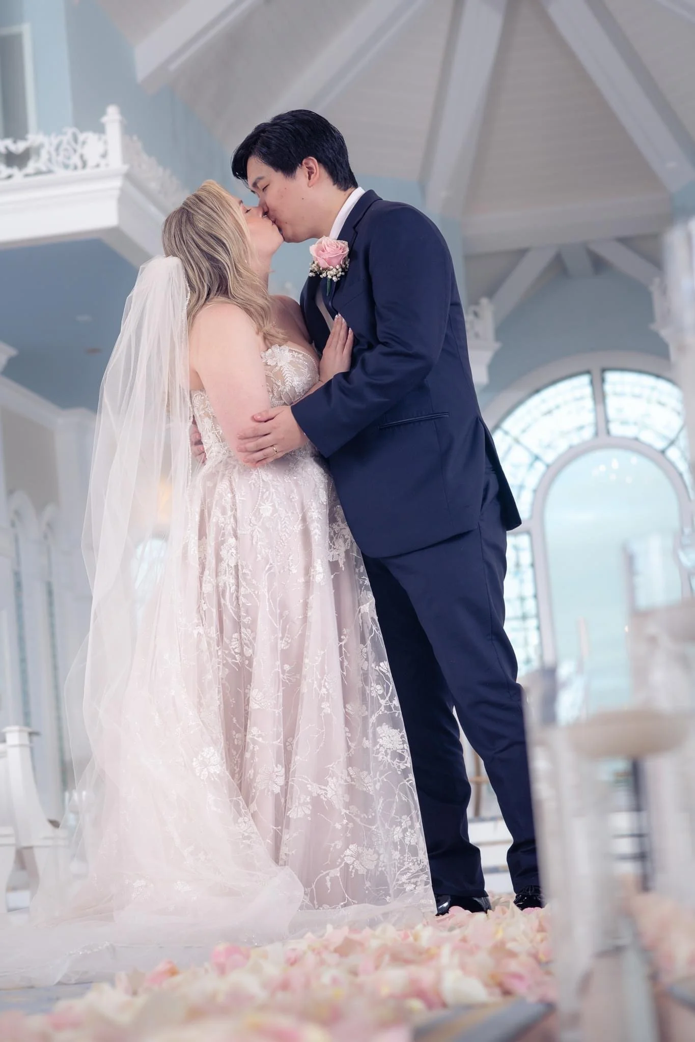 A newlywed couple sharing a kiss during their wedding ceremony, with the bride in a lace wedding dress and veil, and the groom in a navy suit with a pink boutonniere, inside a bright, at Disney's Wedding Pav with large windows and floral decorations 