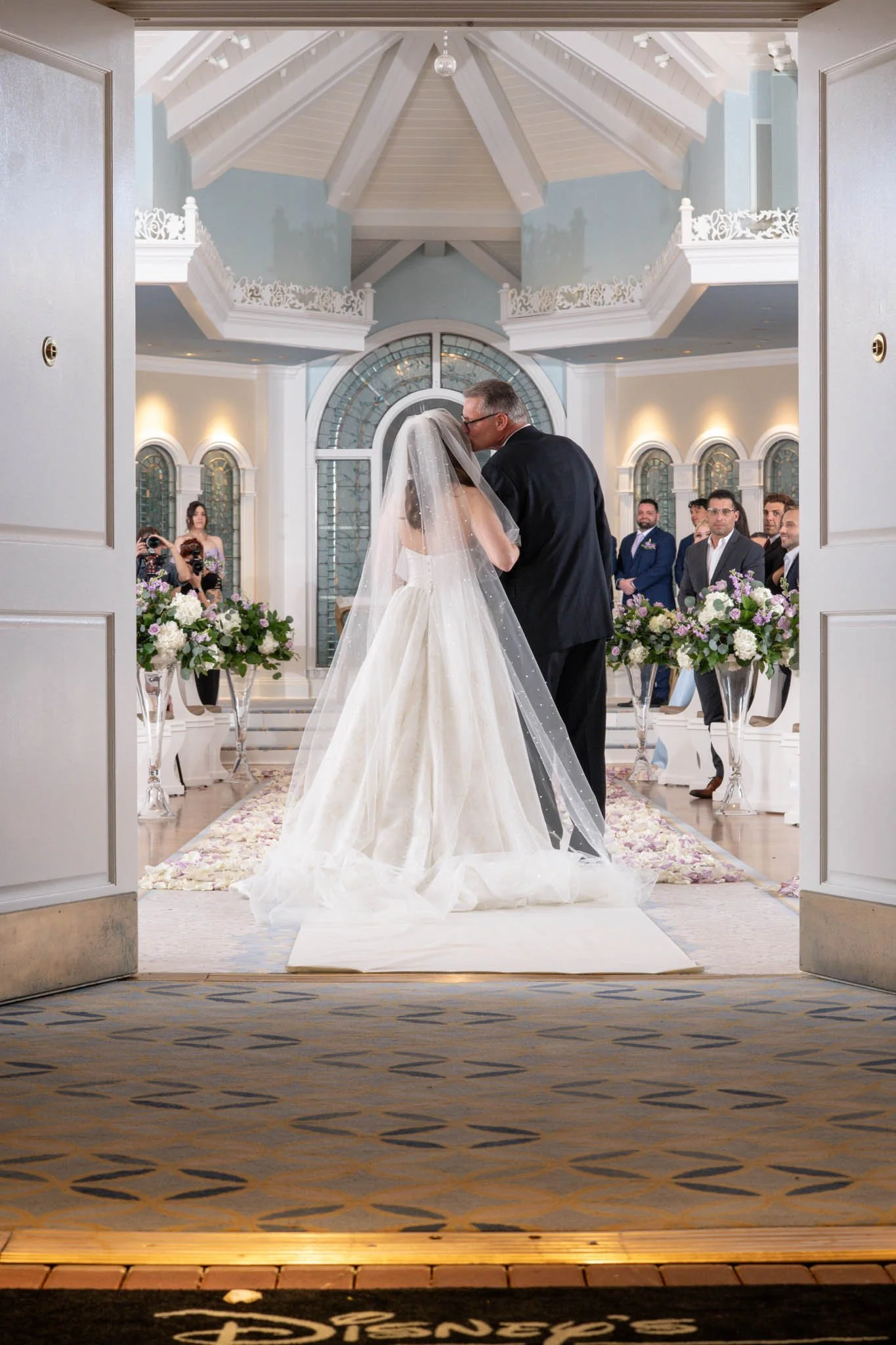 A bride and her father share a moment during a wedding ceremony inside a bright, elegant church with white walls, arched windows, and floral decorations. The bride is wearing a white wedding gown with a long veil, and the father is dressed in a black