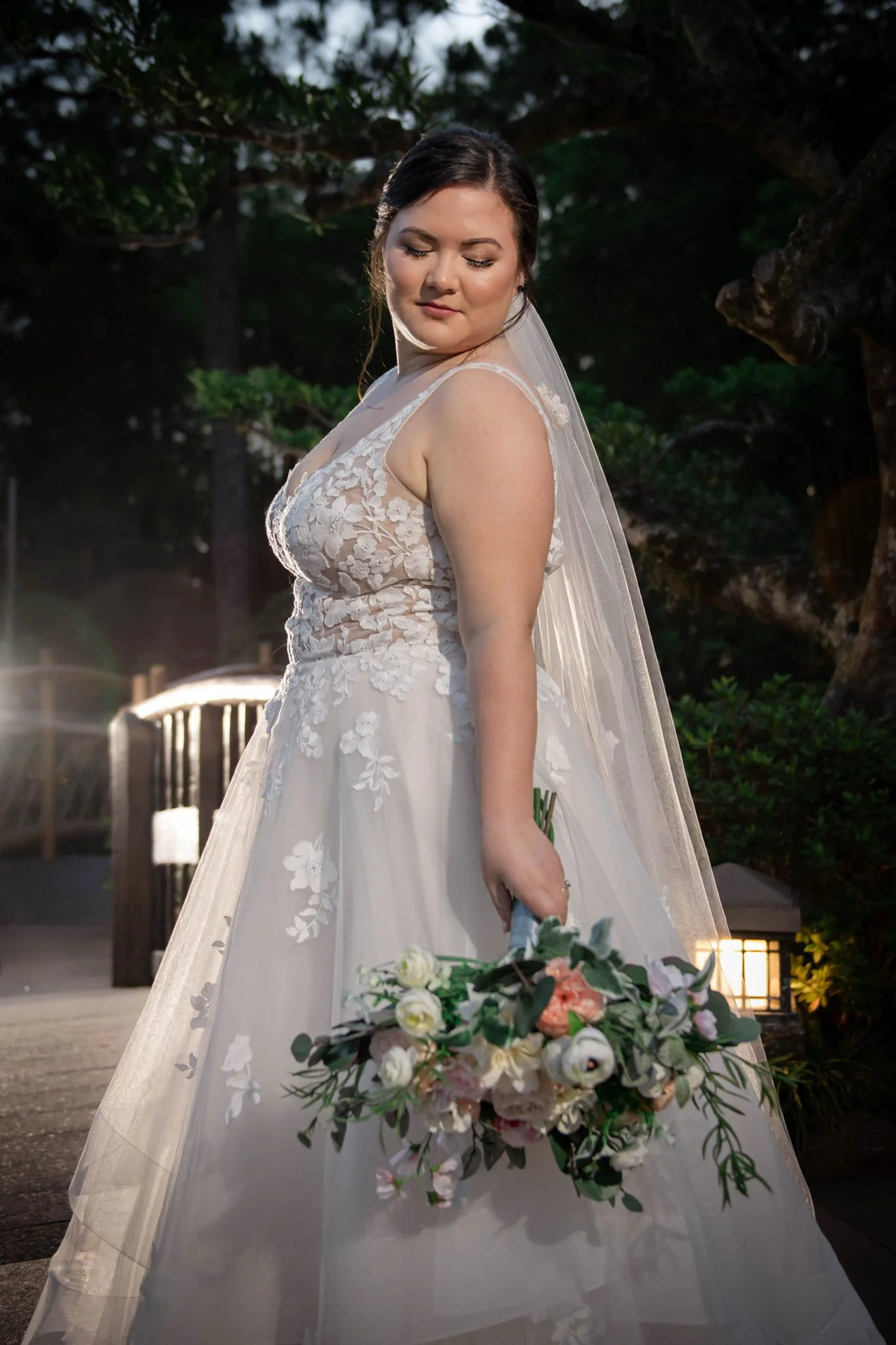 A bride in a white wedding dress with lace details stands outdoors holding a bouquet of white and light pink flowers.
