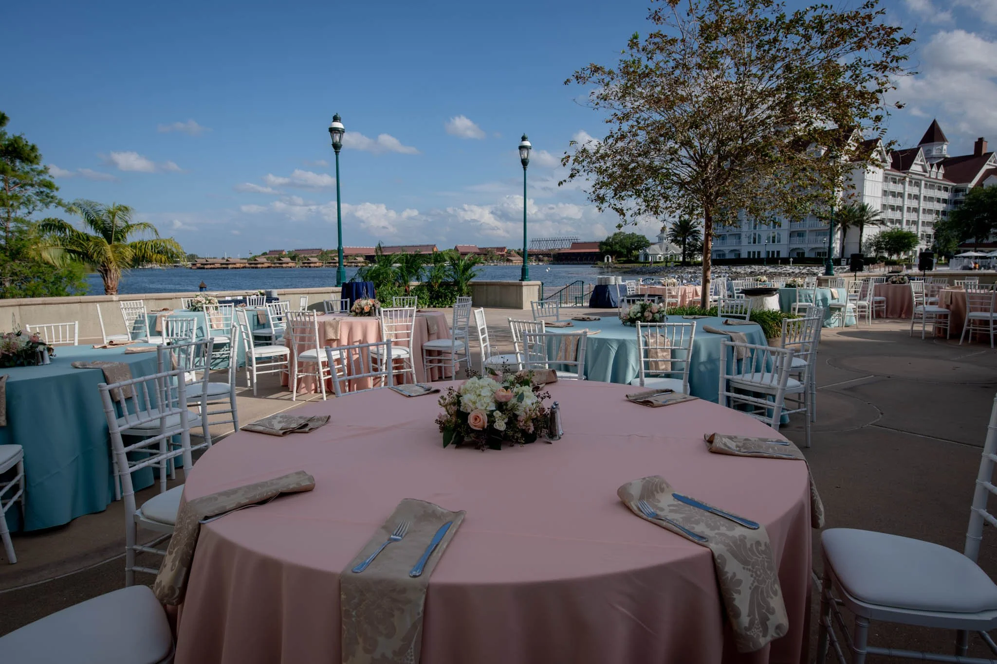Outdoor event space with round tables covered in pastel blue and pink tablecloths, set with silverware and napkins. Centerpieces with flowers are on each table. There are white chairs around the tables, a few trees, and a view of the water with a lar