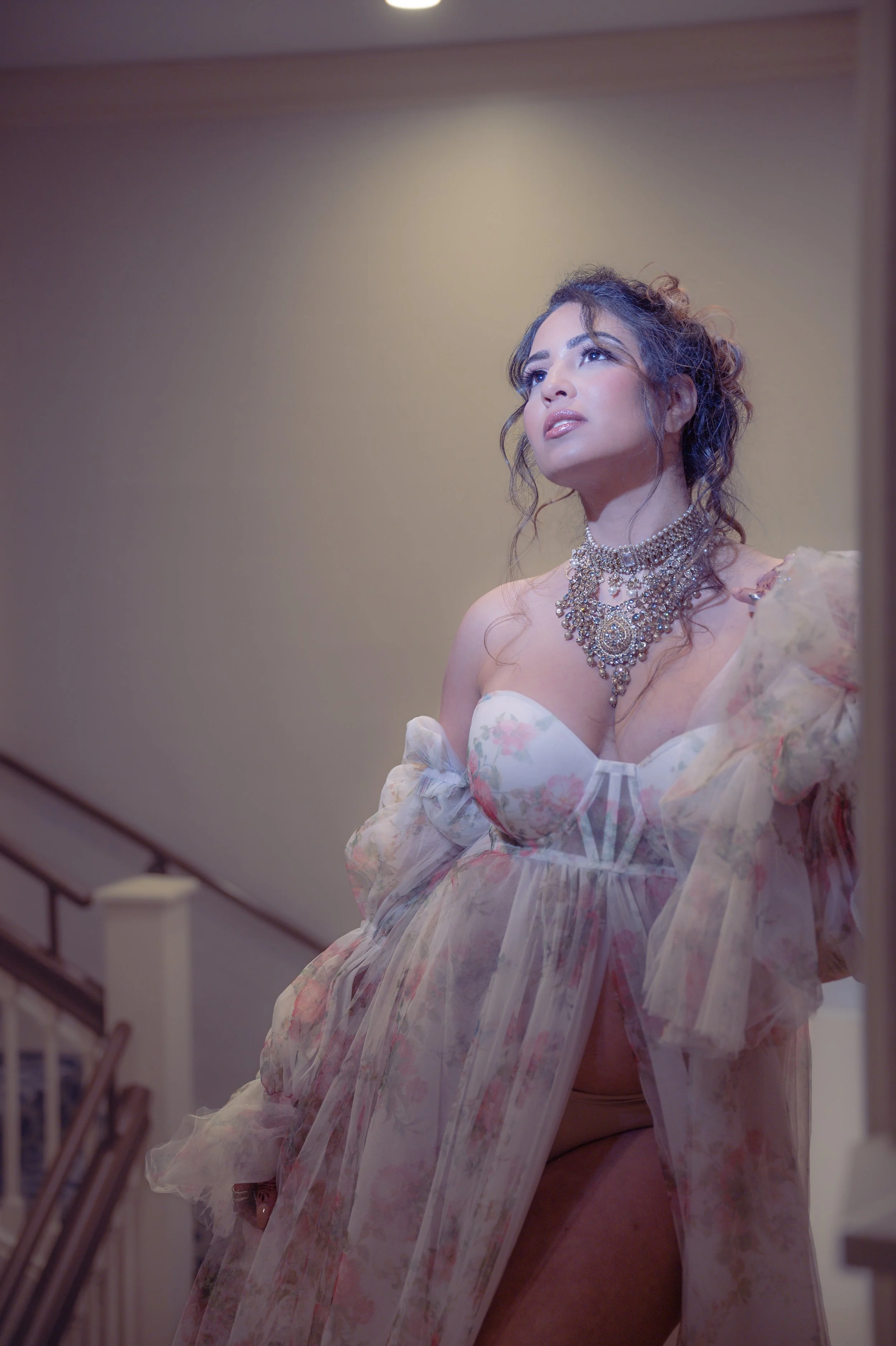 A woman with curly hair looking up, wearing a floral dress with a transparent cape and elaborate jewelry, standing near a staircase.