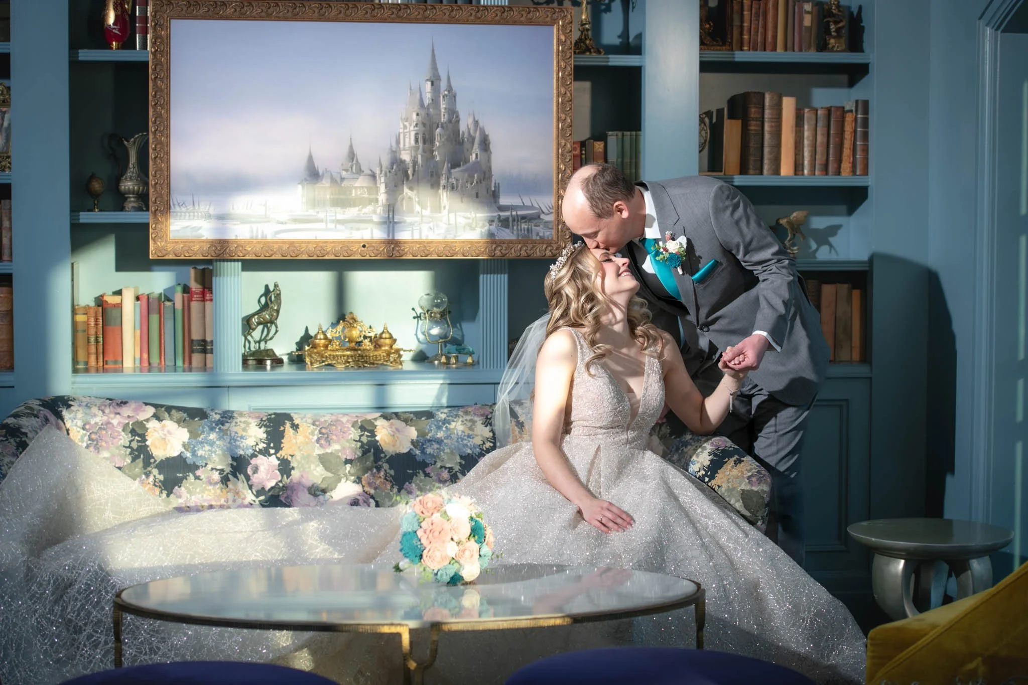 A bride sitting on a floral sofa, wearing a wedding dress and a veil, holding a bouquet, as a groom leans over to kiss her on the forehead in a room with teal walls and bookshelves, a large painting of a castle  above inside the Enchanted Rose.