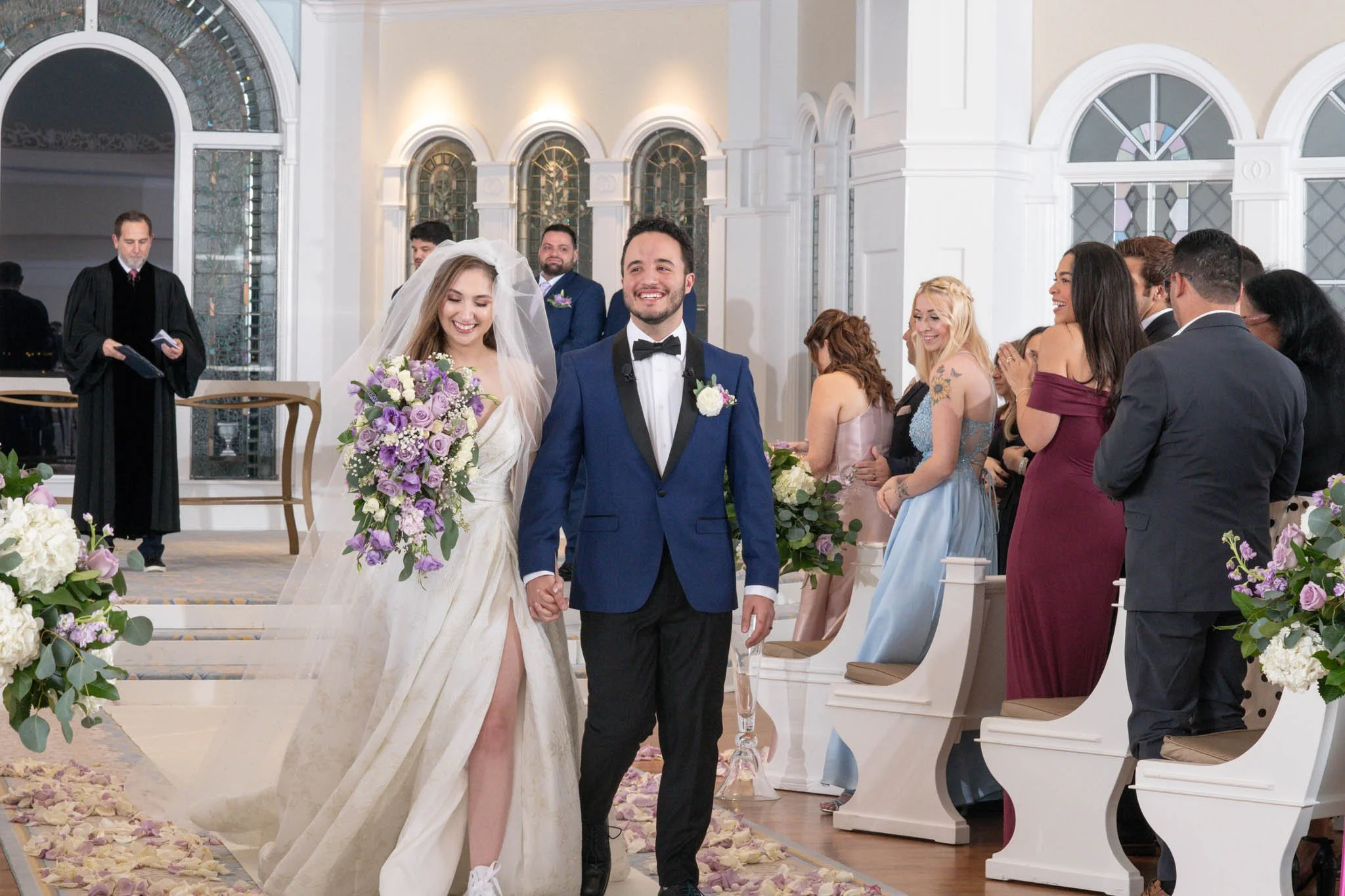 A newlywed couple walking down the aisle, holding hands, while smiling. The bride is dressed in a white wedding gown with a veil and a large bouquet of purple and white flowers. The groom is in a blue tuxedo with a black bow tie and white boutonniere