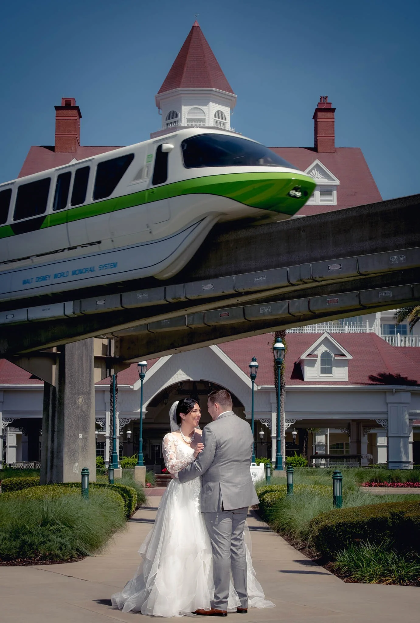 A bride and groom dancing outdoors under a monorail track with a green and white monorail passing overhead, and Disney Grand Floridian with a red roof and spires in the background.