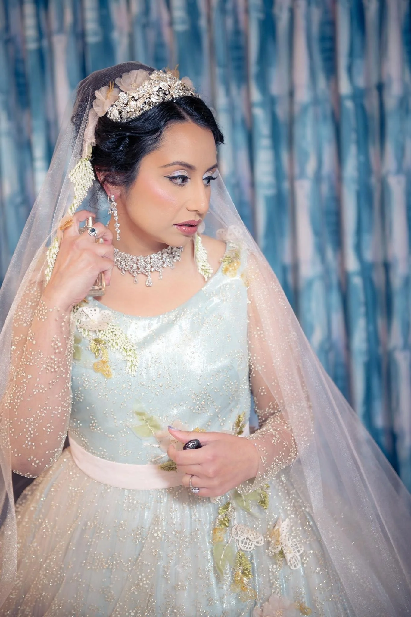 A woman dressed as a bride in a pastel-colored gown with embroidery, wearing a tiara, a necklace, earrings, and a veil, holding a perfume bottle, with a semi-abstract blue backdrop.