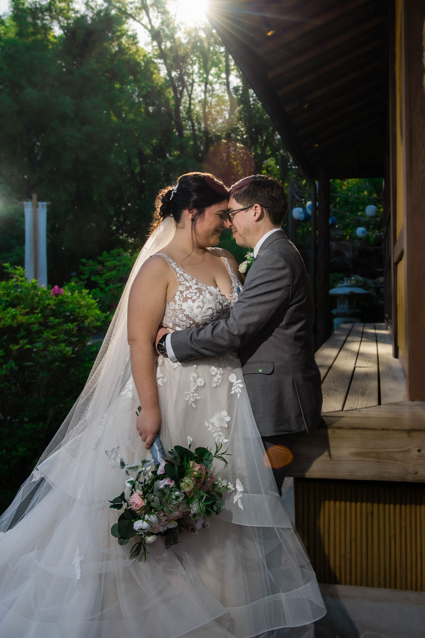 A bride and groom share an intimate moment outdoors during sunset, with noses touching and eyes closed, surrounded by greenery, sunlight, and a wooden structure.