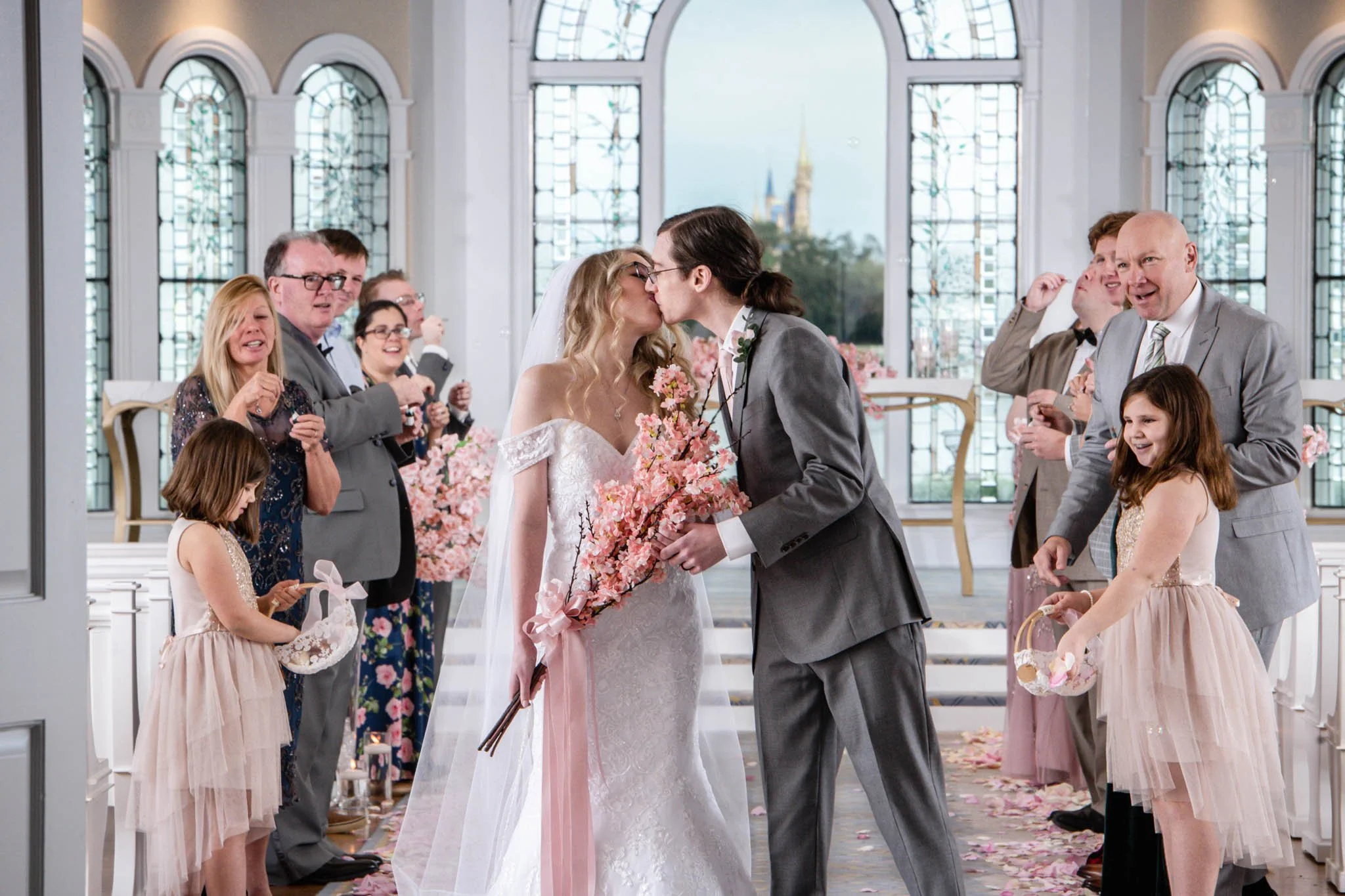A wedding ceremony at Disney's Wedding Pavilion with a bride and groom kissing, surrounded by guests inside a decorated venue with large arched windows and pink flower decorations.