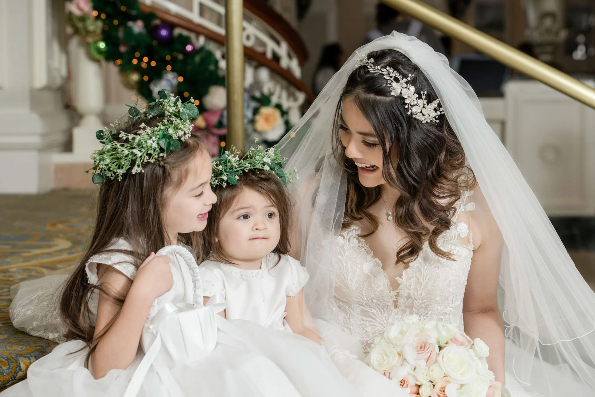 Bride in wedding dress with dark wavy hair and veil, smiling and talking to three young girls in white dresses with floral crowns, in a decorated indoor setting with a Christmas tree in the background.