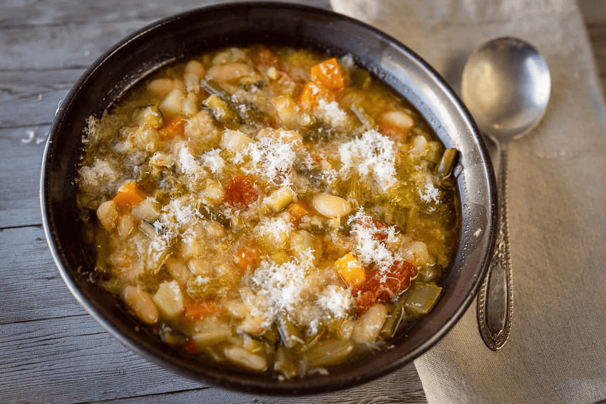 A bowl of vegetable and bean soup topped with grated cheese, on a wooden table with a spoon and napkin.