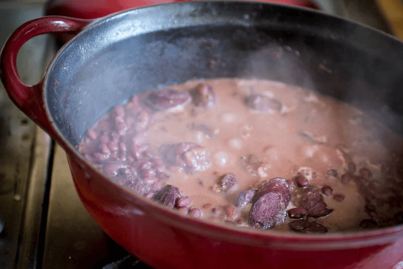A red saucepan on a stove with boiling meat and broth inside.