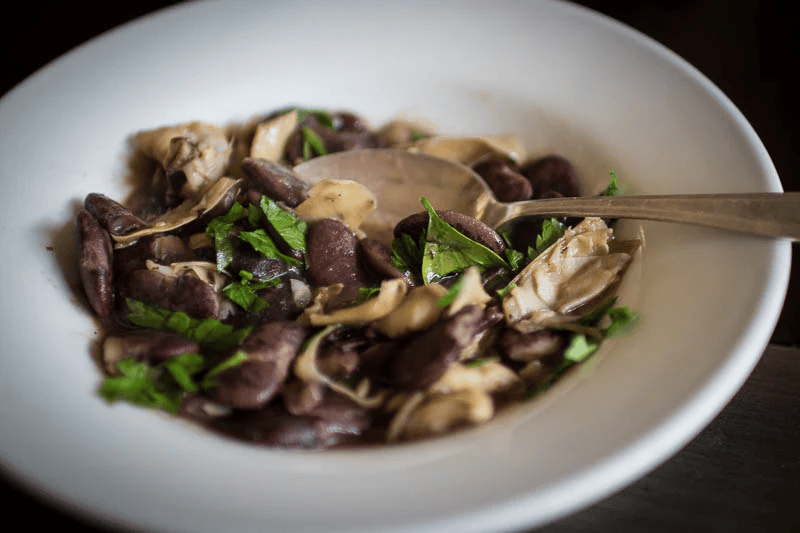 A bowl of cooked purple and beige mushrooms garnished with chopped green herbs and a spoon inside.