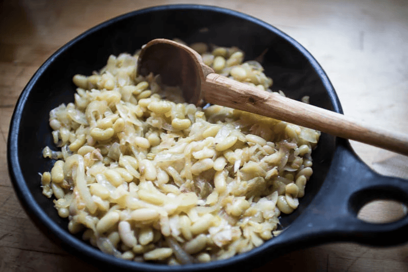 A black skillet with cooked white beans and onions, and a wooden spoon resting inside. The skillet is placed on a wooden surface.