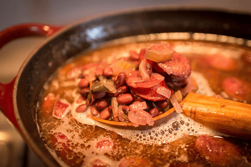 Close-up of a skillet filled with cooked beans, sausage slices, and vegetables, with a wooden spoon resting inside.