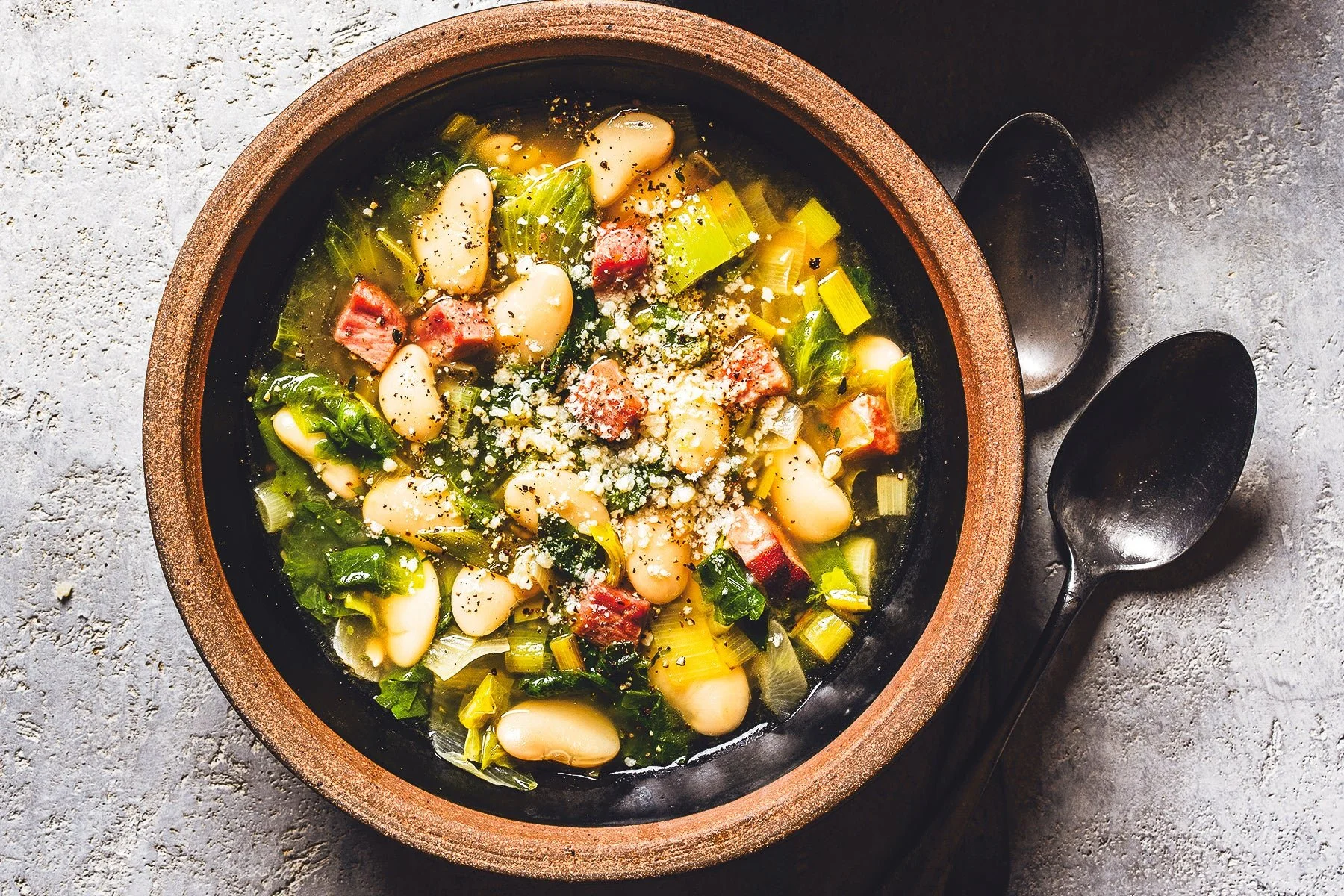 Soup with beans, celery, bacon, and cheese in a bowl on a gray surface with two black spoons beside it.