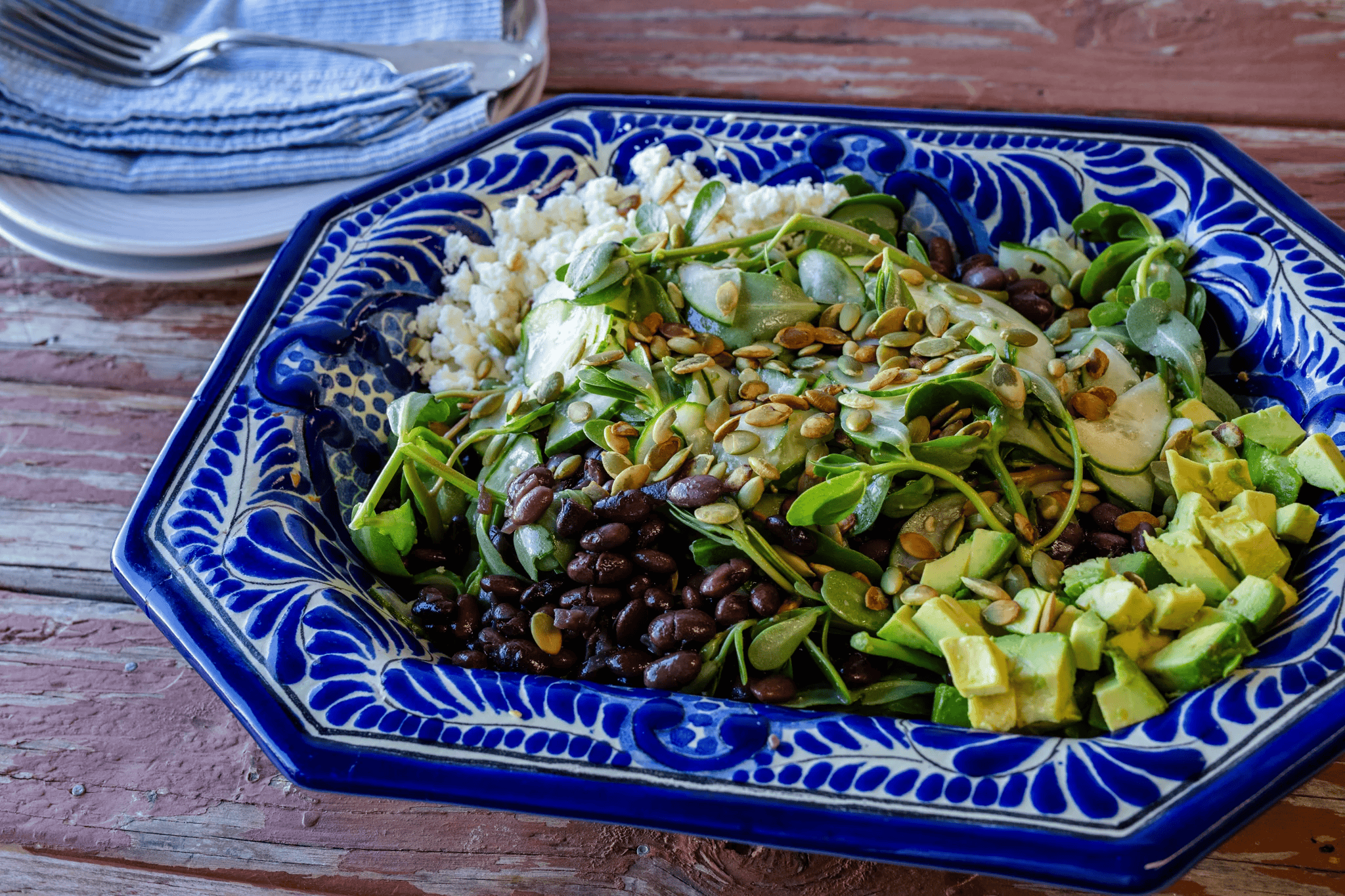 A colorful salad in a decorative blue and white bowl containing black beans, sliced cucumber, avocado, sunflower seeds, and crushed feta cheese, placed on a rustic wooden table.