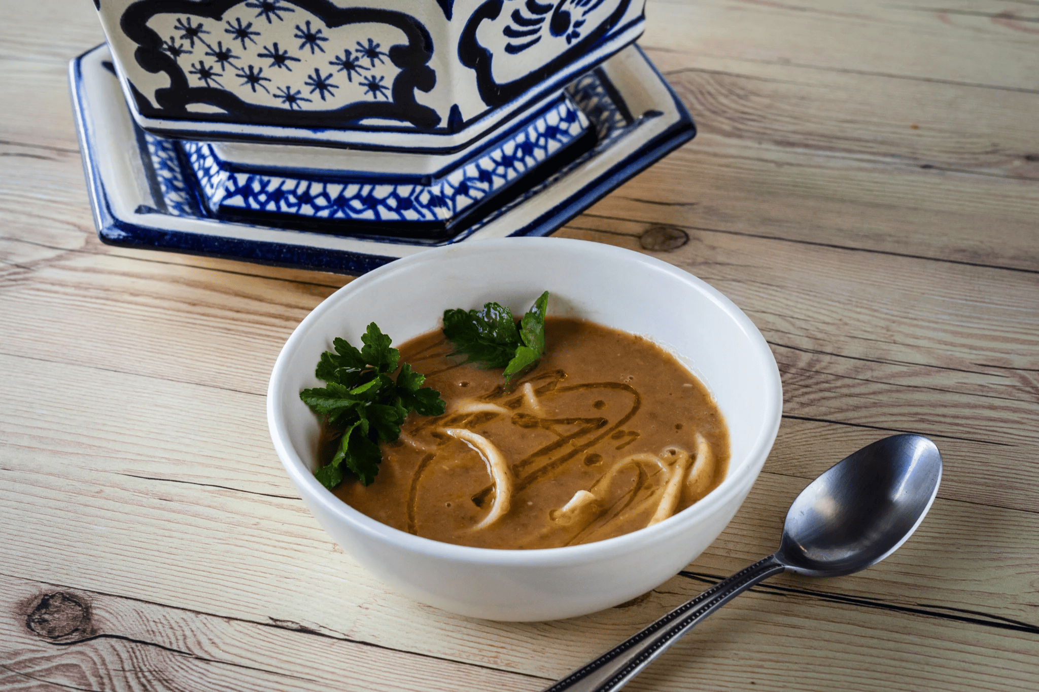 Bowl of creamy brown soup garnished with fresh parsley on a wooden table, with a silver spoon nearby and decorative blue and white patterned ceramic dishes in the background.