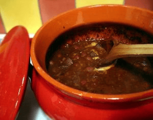A red ceramic pot filled with dark, thick soup or stew, with a wooden spoon resting inside.