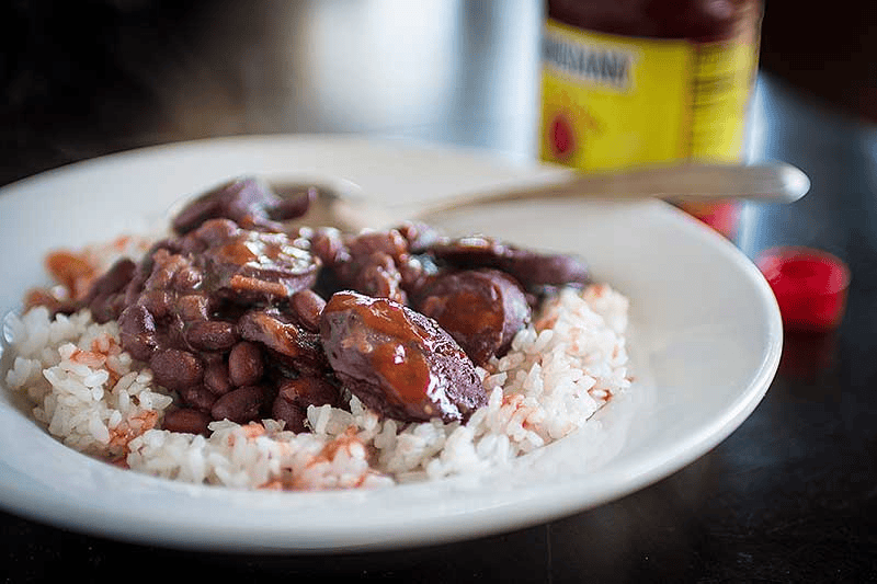 A bowl of white rice topped with dark red chili beans in a thick sauce, accompanied by a spoon, with a can of chili and a small red chili pepper in the background.