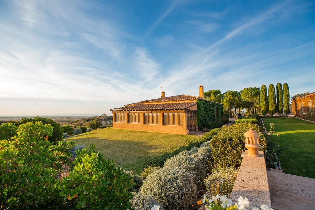 A large house with a terracotta roof and a sunroom, surrounded by lush green landscaping, tall cypress trees, and a clear blue sky at sunset.