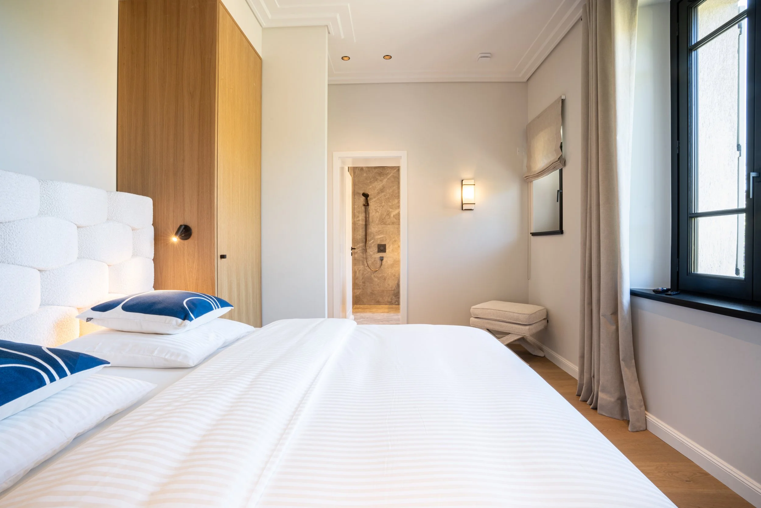 Chambre moderne avec un lit fait avec des oreillers bleus et blancs, un mur en bois, une fenêtre avec rideau beige, et une salle de douche visible en arrière-plan.