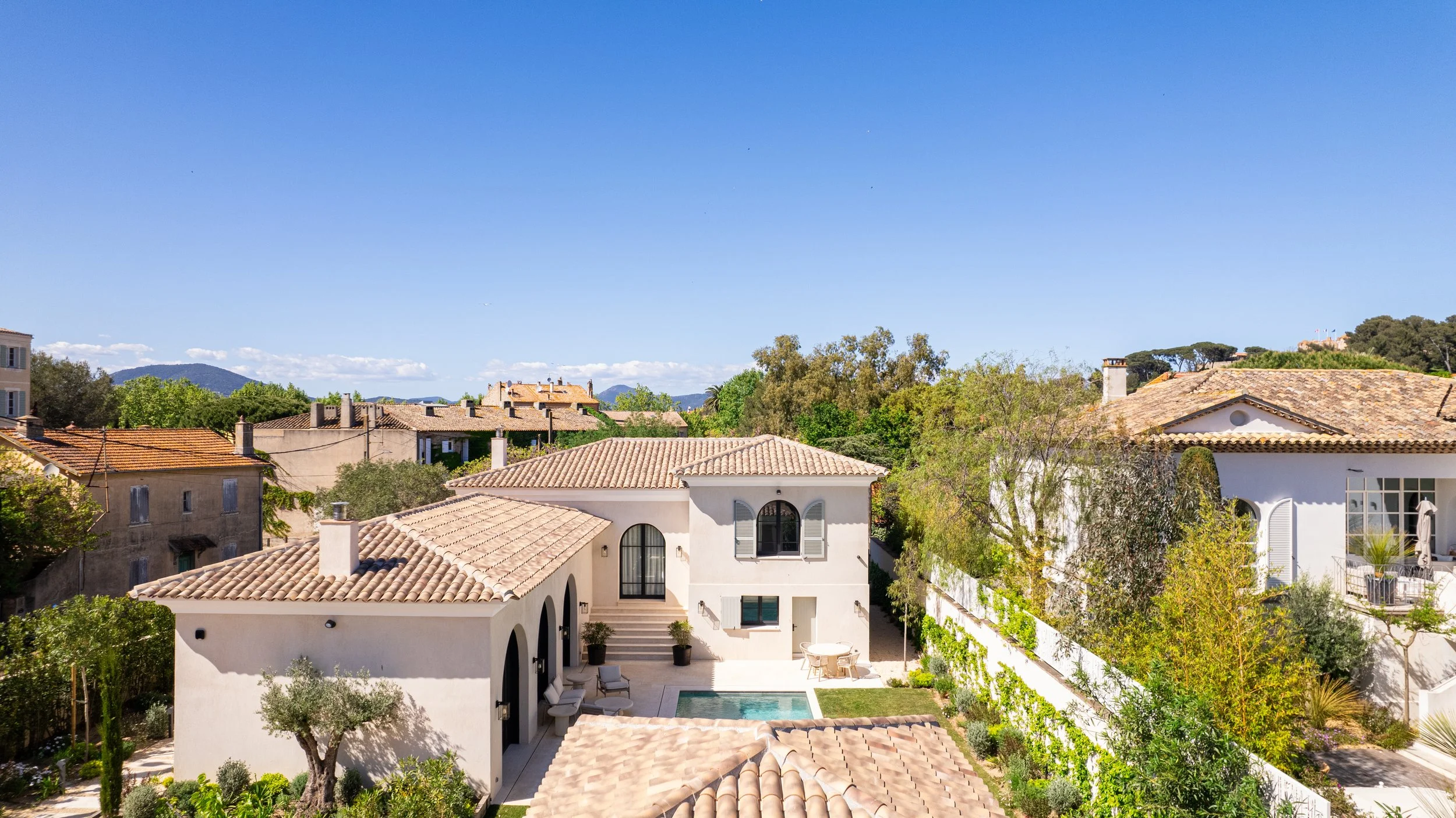 Maisons de style méditerranéen avec toits en tuiles, jardin avec arbres, piscine, sous un ciel bleu clair.
