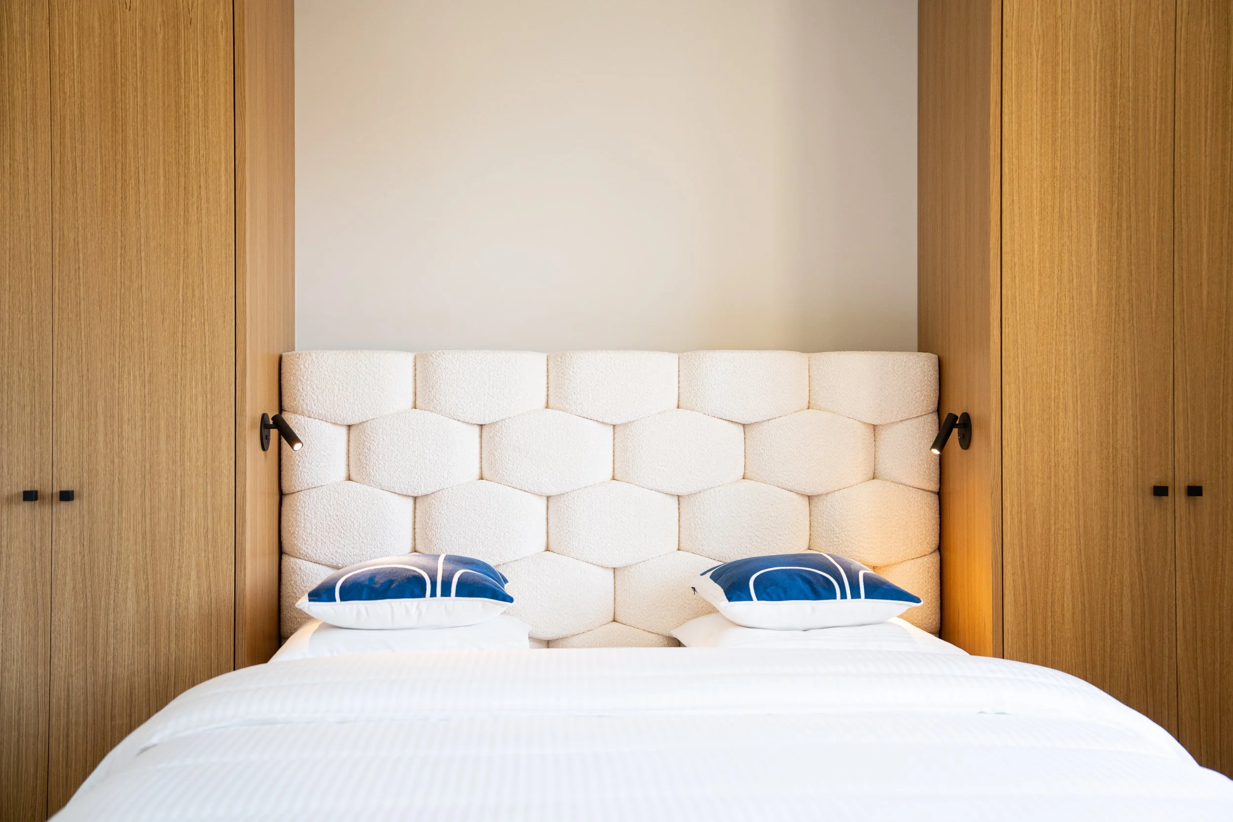 Lit avec une tête de lit en tissu beige, deux lampes de lecture, et deux oreillers bleus avec motif blanc, encadré par des armoires en bois.