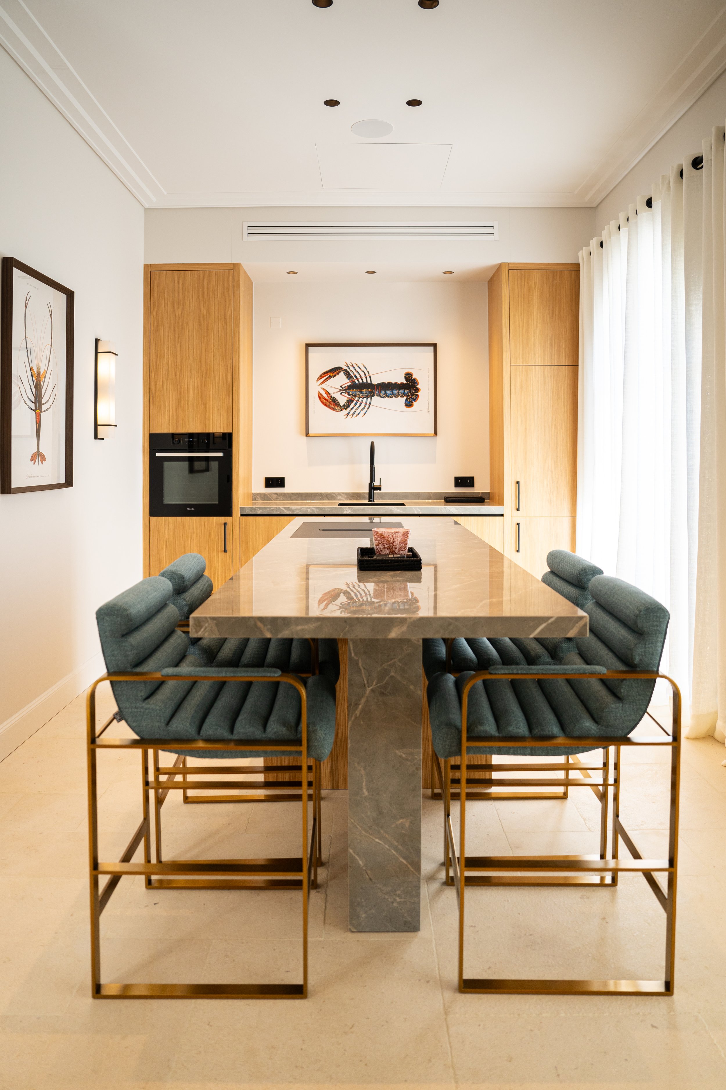 Cuisine moderne avec un îlot en marble, fauteuils de salle à manger en tissu vert et métal doré, murs blancs, et une œuvre d'art représentant un homard au mur.