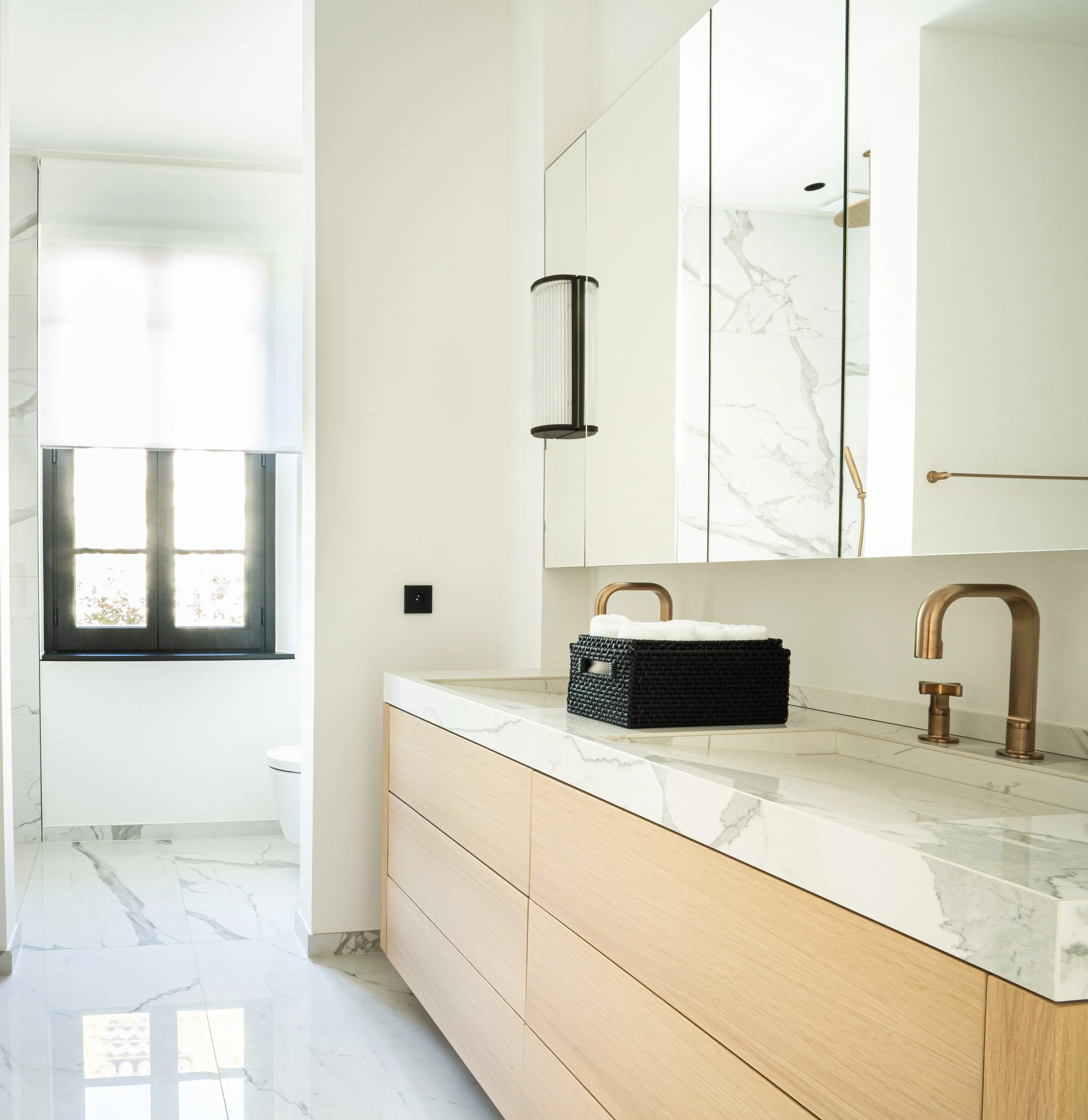 Salle de bain moderne avec un meuble en bois clair, un plan de travail en marbre blanc avec des veines grises, un miroir mural, une robinetterie en bronze, et une fenêtre laissant entrer la lumière naturelle.