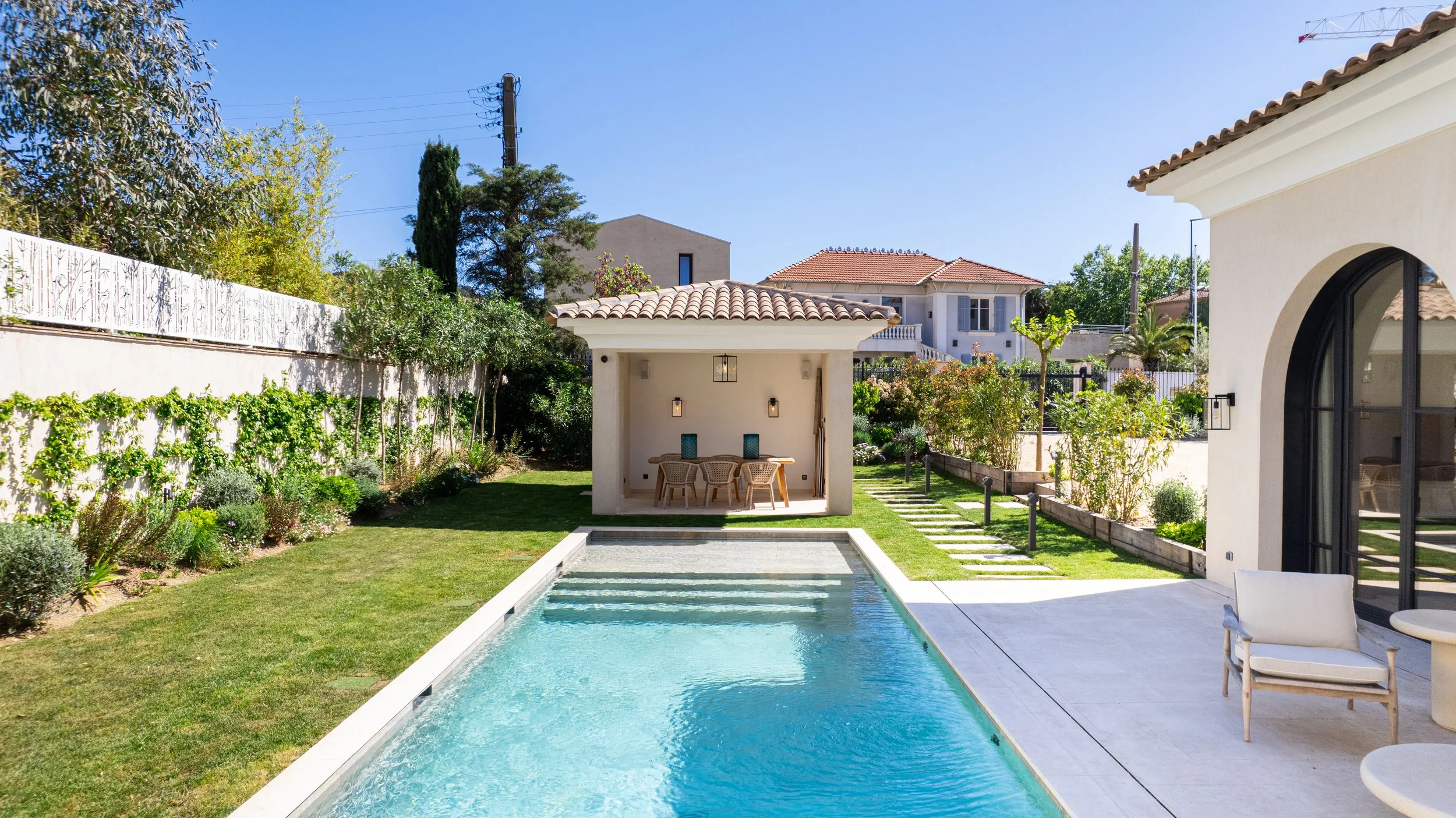Piscine dans un jardin ensoleillé avec une maison moderne à droite, un espace terrasse avec des fauteuils, et un petit cabanon avec table et chaises au fond, entouré de végétation et de plantes