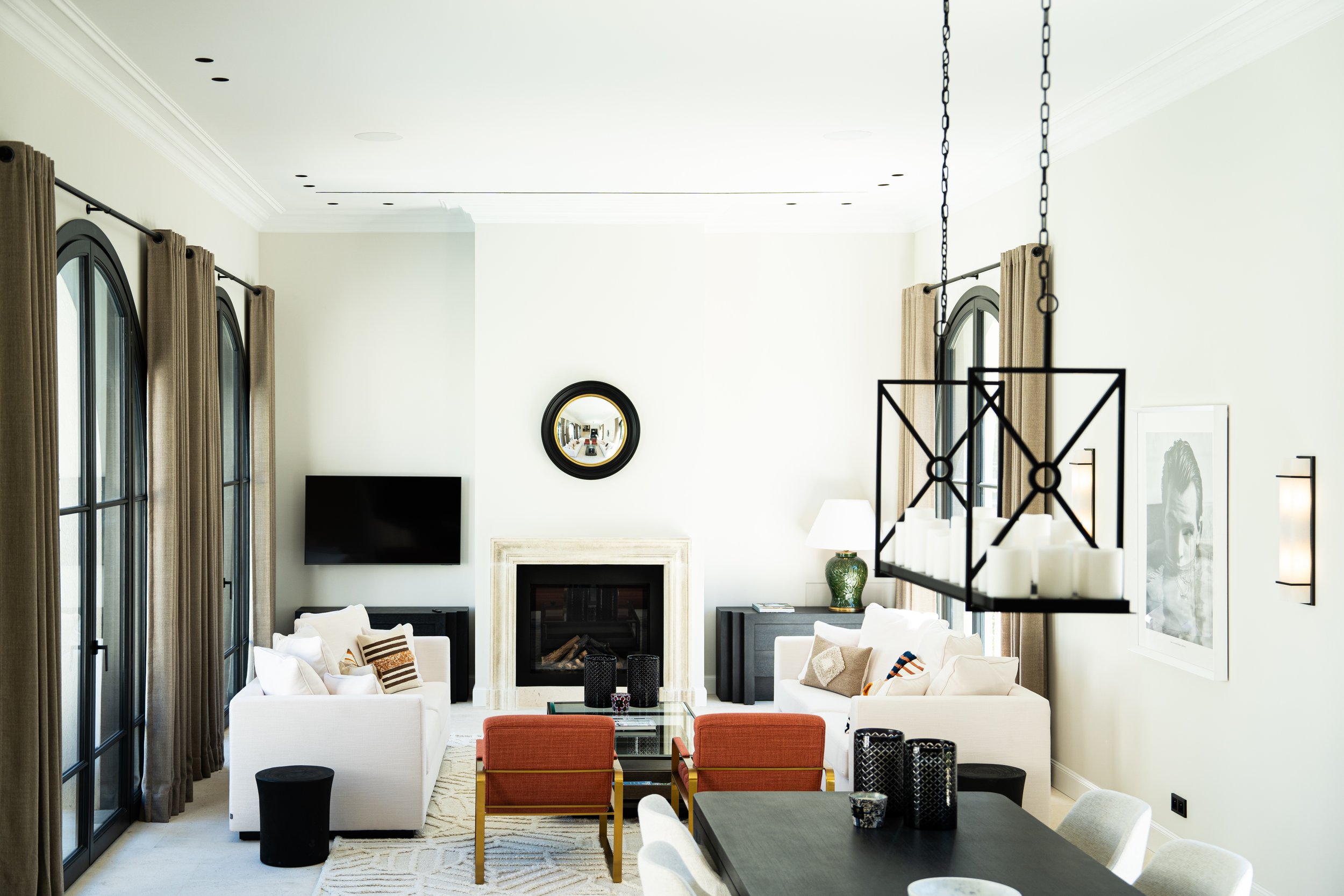 Salon moderne avec cheminée, canapés blancs, table basse en verre, fauteuils orange, lampes sur tables, grand tapis, mur blanc avec miroir rond et peinture encadrée, téléviseur mural, fenêtres avec rideaux marron.