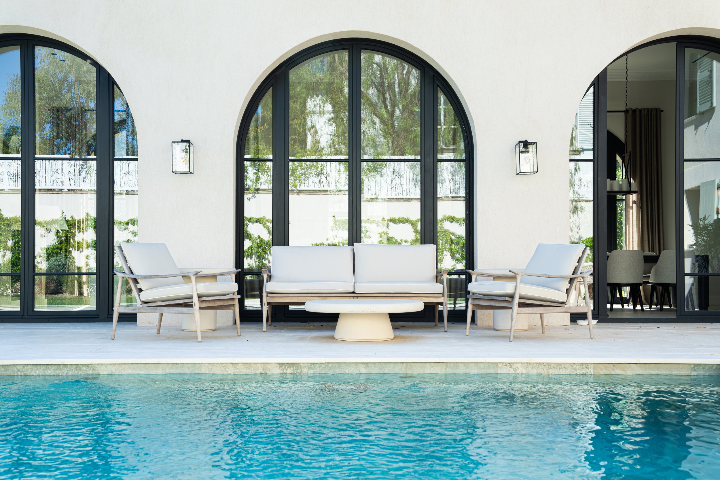 Façade d'une maison avec grandes fenêtres arquées, mobilier de salon beige à l'extérieur et vue sur la piscine.
