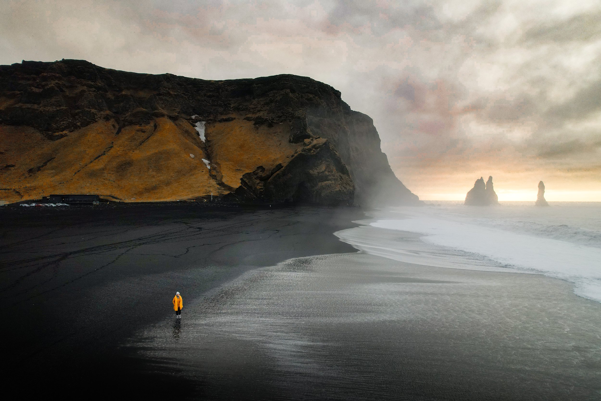 Une plage noire avec une falaise escarpée en arrière-plan, deux formations rocheuses dans la mer au loin, et une personne portant une veste jaune marchant seul sur la plage par temps nuageux.