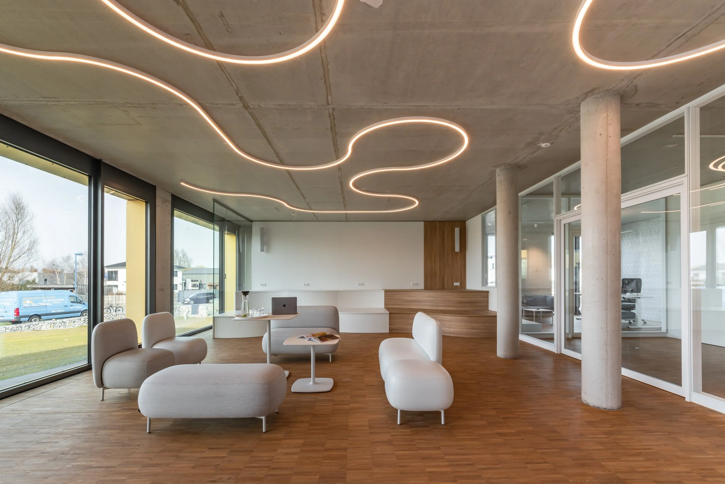 Moderne, leere Lounge mit weißen Sitzmöbeln, großen Fenstern, Holzfußboden, Deckenleuchten in Wellenform, und Glaswänden, die einen Büroraum abtrennen.