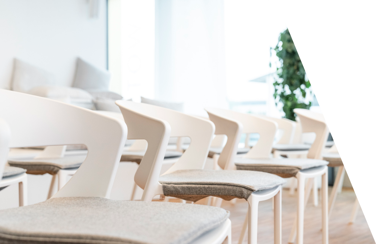Leerer Seminarraum mit weißen Stühlen und grauen Sitzkissen, große Fenster mit Blick nach außen, Pflanzen im Hintergrund.