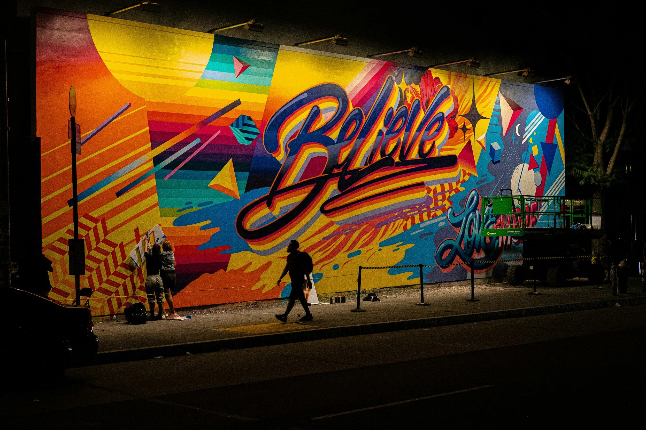 Nighttime view of a large colorful mural being painted on an exterior wall, with people working on it and a lift parked nearby.