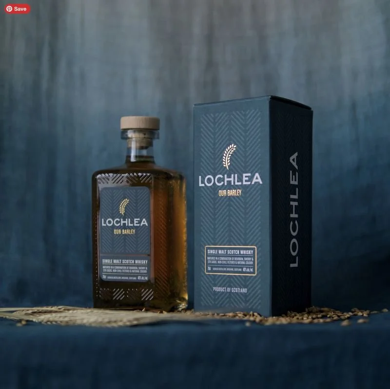 A bottle of Lochlea single malt Scotch whiskey next to its packaging box on a dark surface, with scattered grains and a blue textured background.