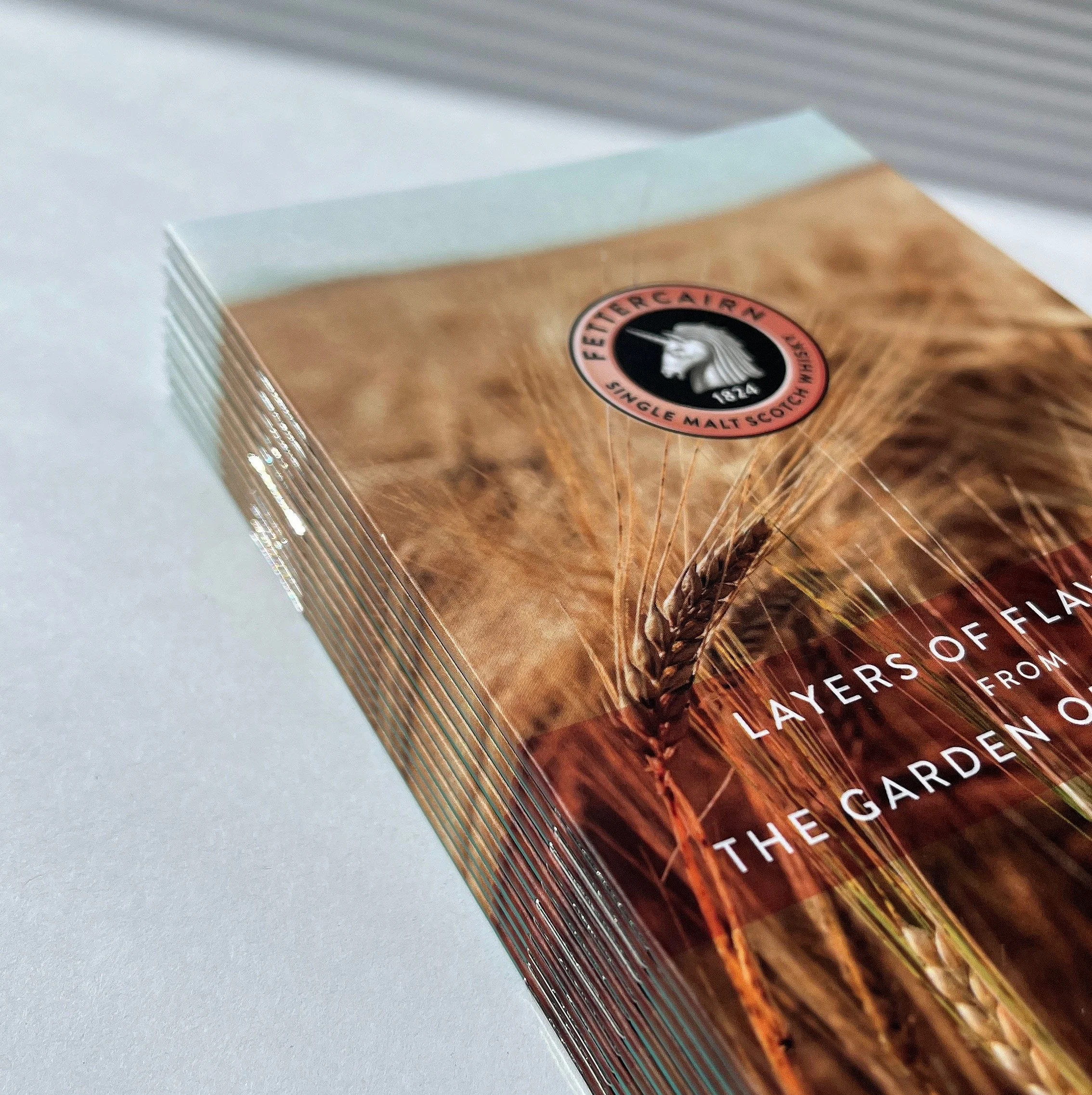 Close-up of a brochure or pamphlet featuring wheat stalks and a circular logo that says "Fettercairn Single Malt Scotch Whisky" with a unicorn illustration. The text reads "Layers of Flavor from the Garden of Earth".