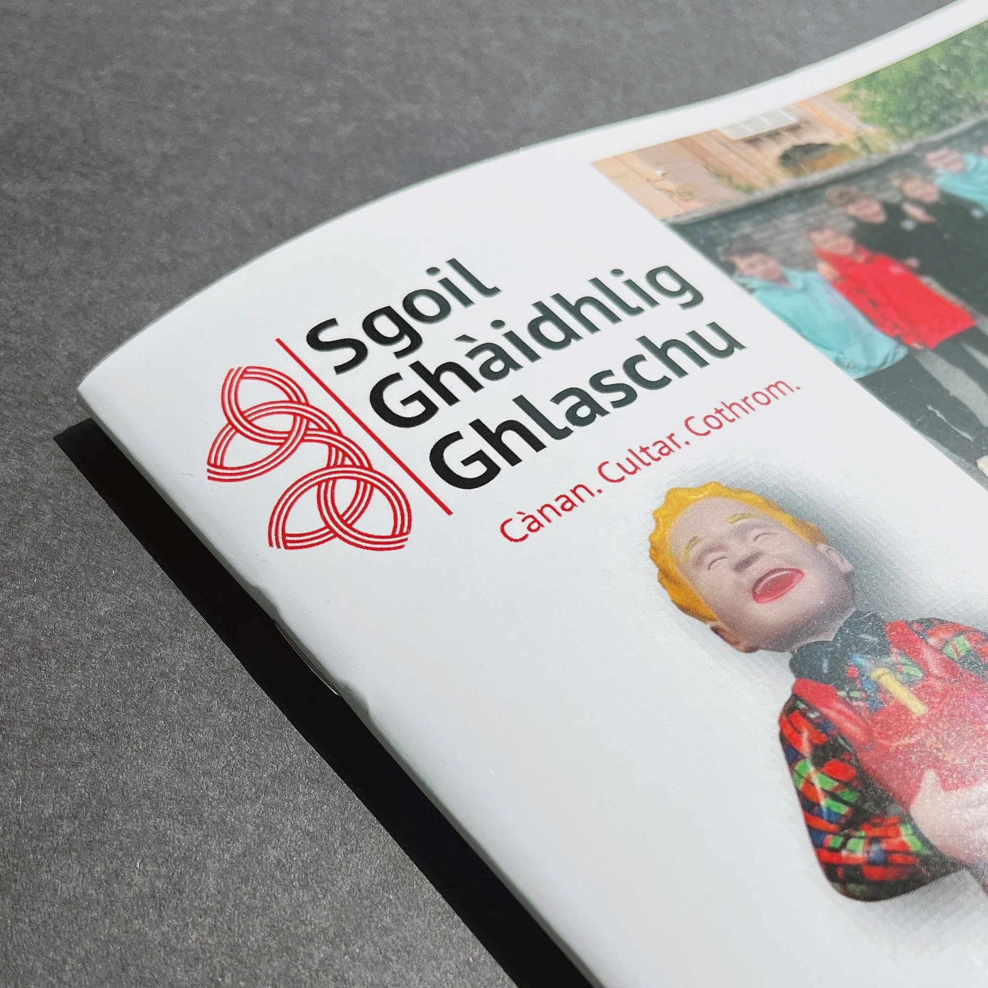 Close-up of a printed publication with the title in Irish, featuring a picture of a girl with blonde hair and bright red lipstick, wearing a colorful jacket. The cover includes red and black text and a decorative red Celtic knot design on a dark grey surface.