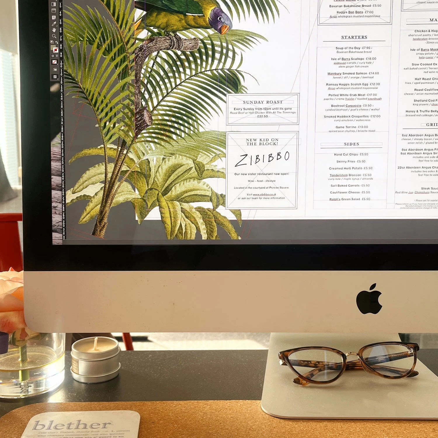 Computer screen displaying a menu with an illustration of tropical leaves and a parrot, glasses resting on the desk to the right side, and a small candle and a flower in a glass vase on the left side.