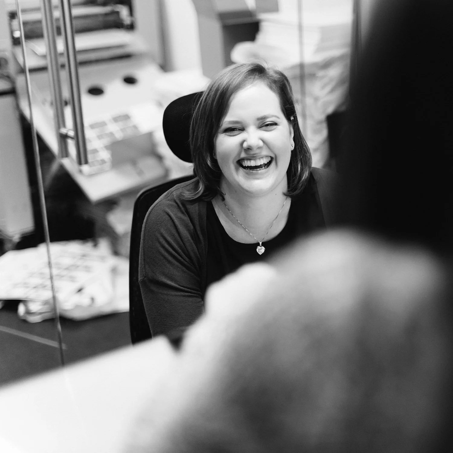 A woman with shoulder-length hair laughing while talking to someone in an office setting.